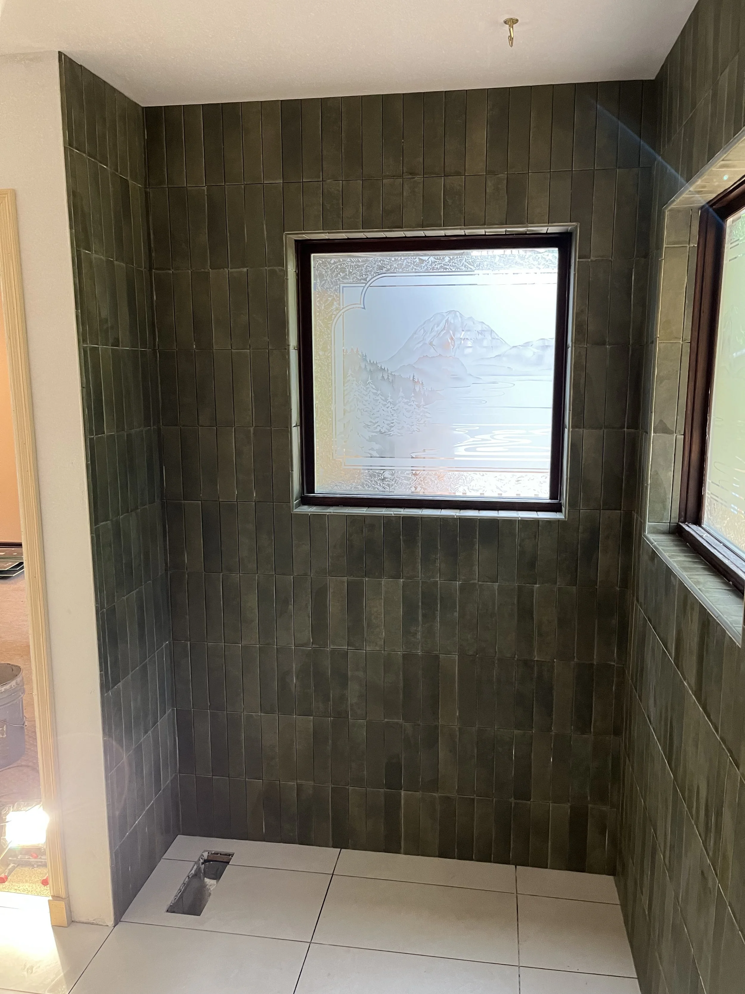 Bathroom with green tiled walls, two windows with frosted mountain landscape glass, and a white tiled floor, with an open plumbing fixture in the floor.