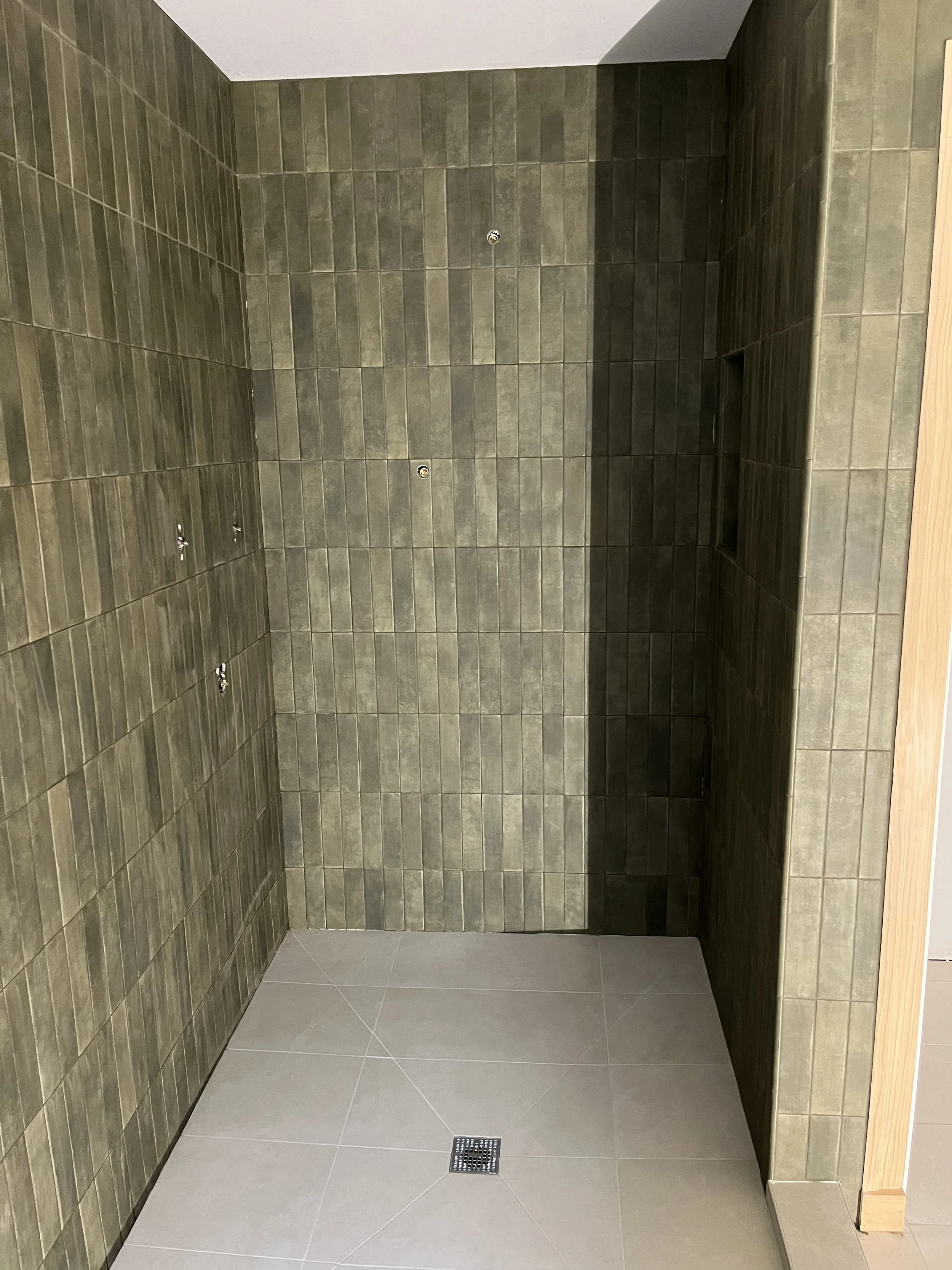 Empty shower stall with green tiled walls, a drain on the floor, and a small shelf for toiletries.