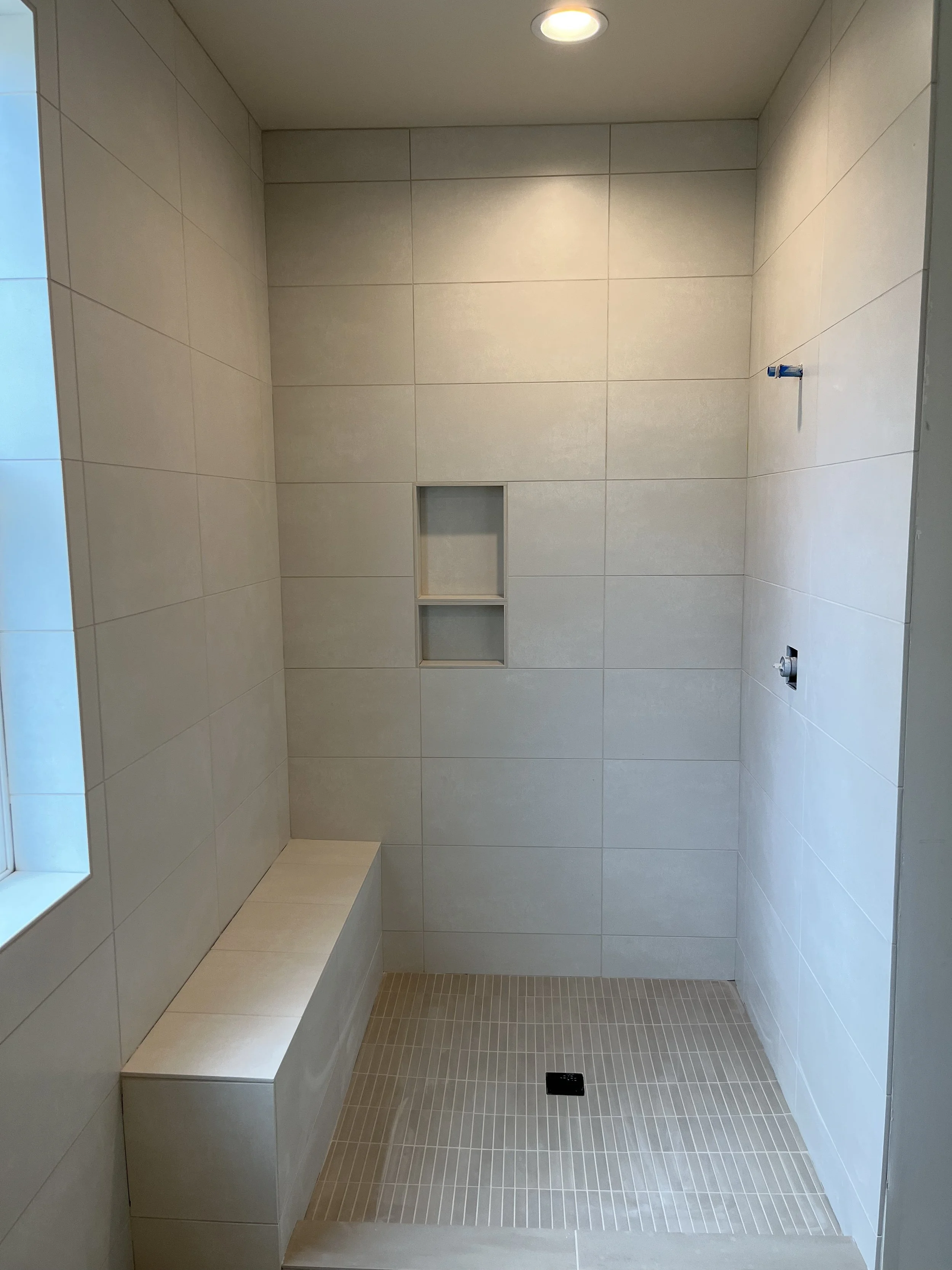 Empty private shower room with beige tiled walls, a built-in bench, a small niche for toiletries, and a floor drain.