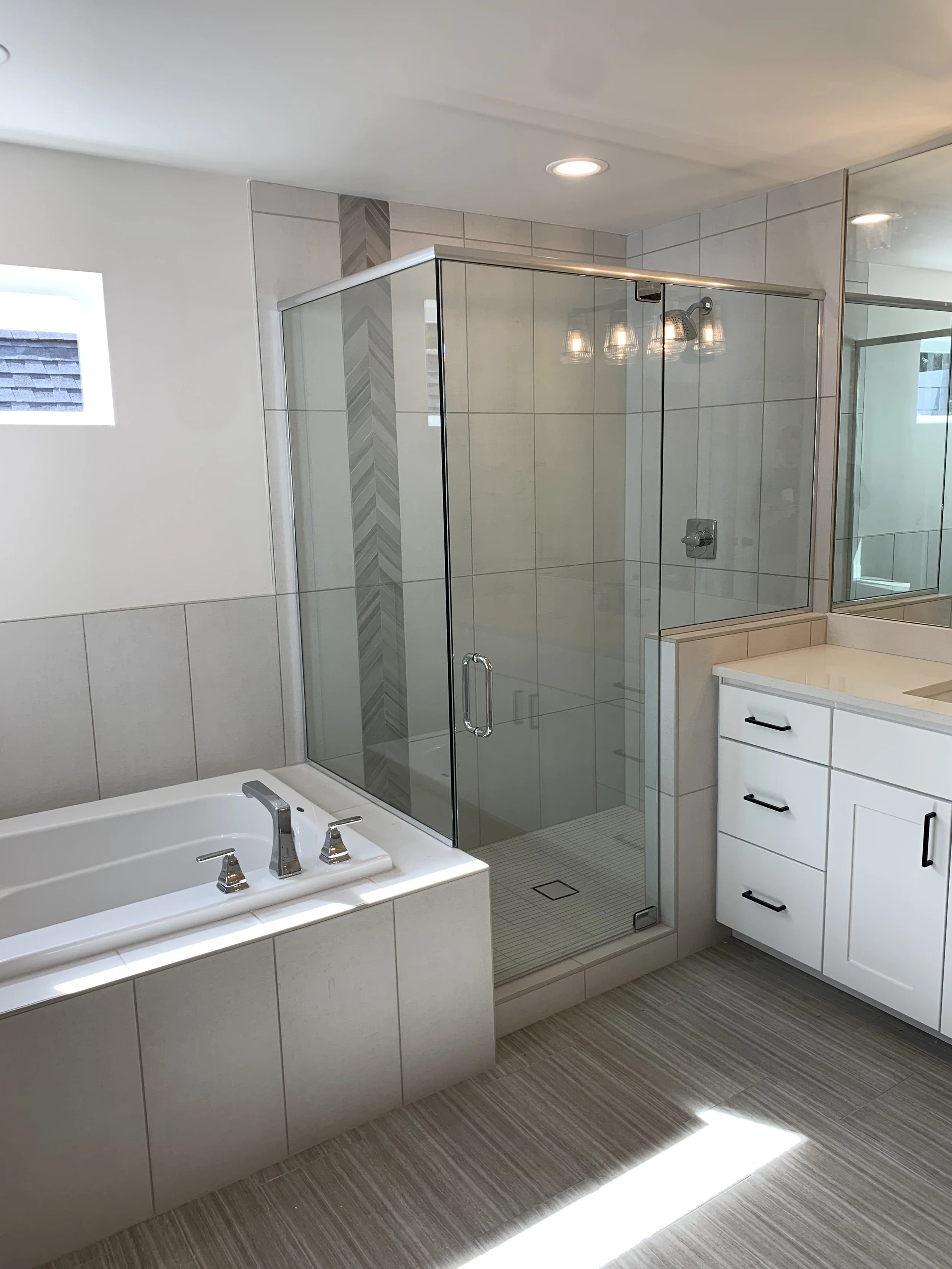 A modern bathroom with a glass-enclosed shower, a bathtub, and a white vanity with black handles. The room has tiled walls and floors, a small window, and recessed ceiling lights.