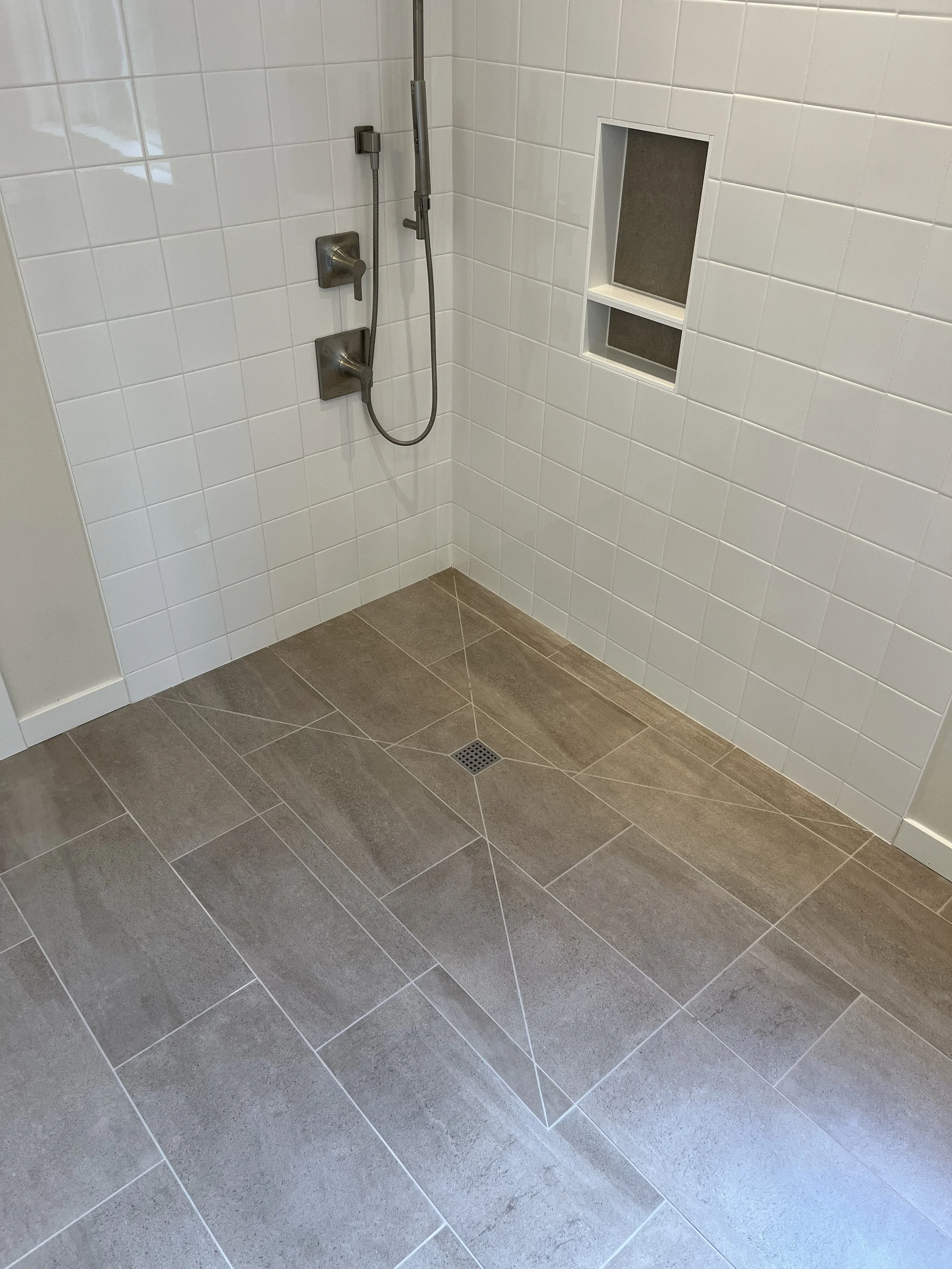 Shower area with white tiled walls, a built-in shelf, and grey tiled floor, featuring a showerhead, controls, and a drain.