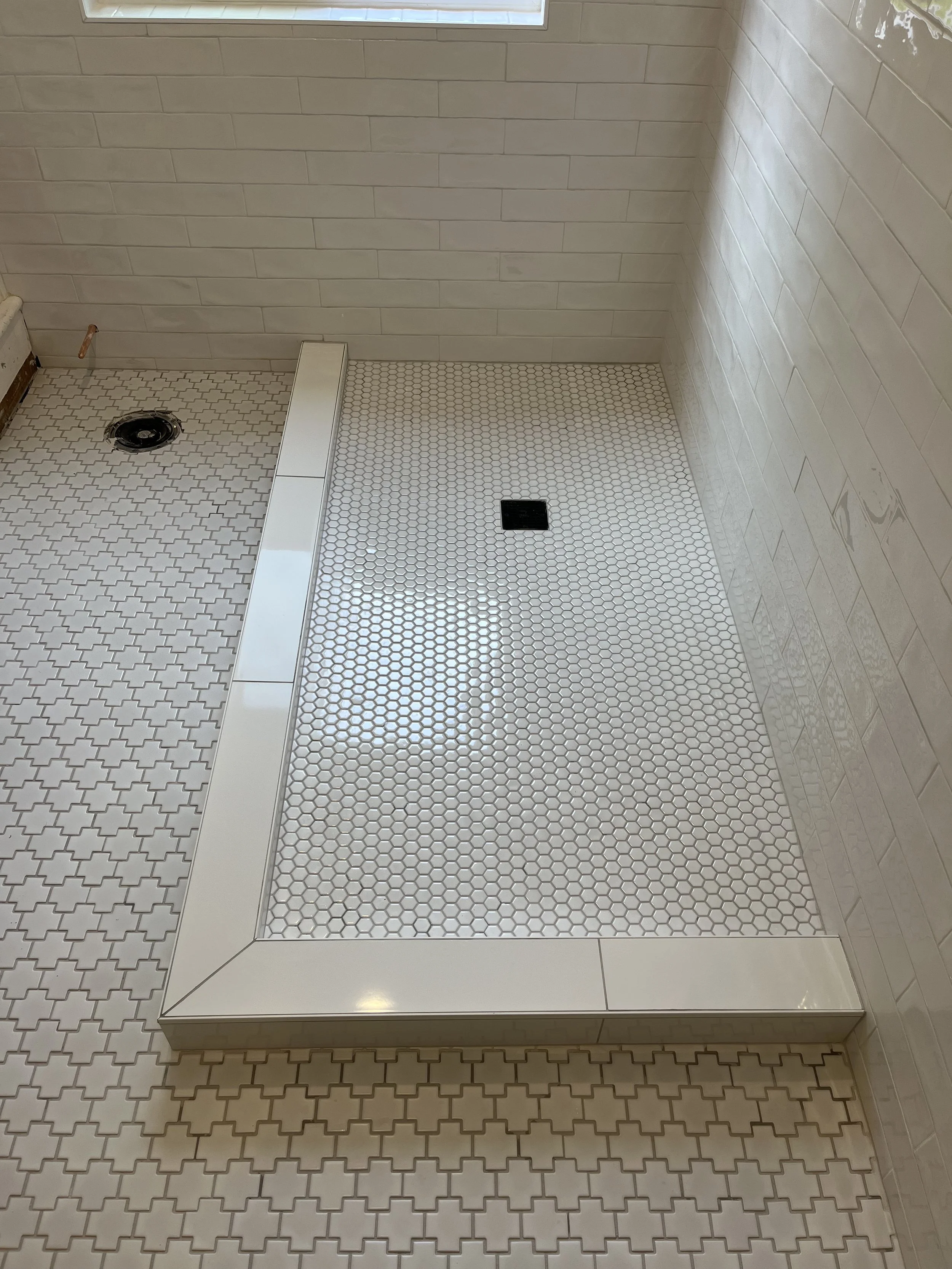 Bathroom shower area with white hexagon tiles, a drain, a wall drain, and beige subway tiles on the walls, with a window above.