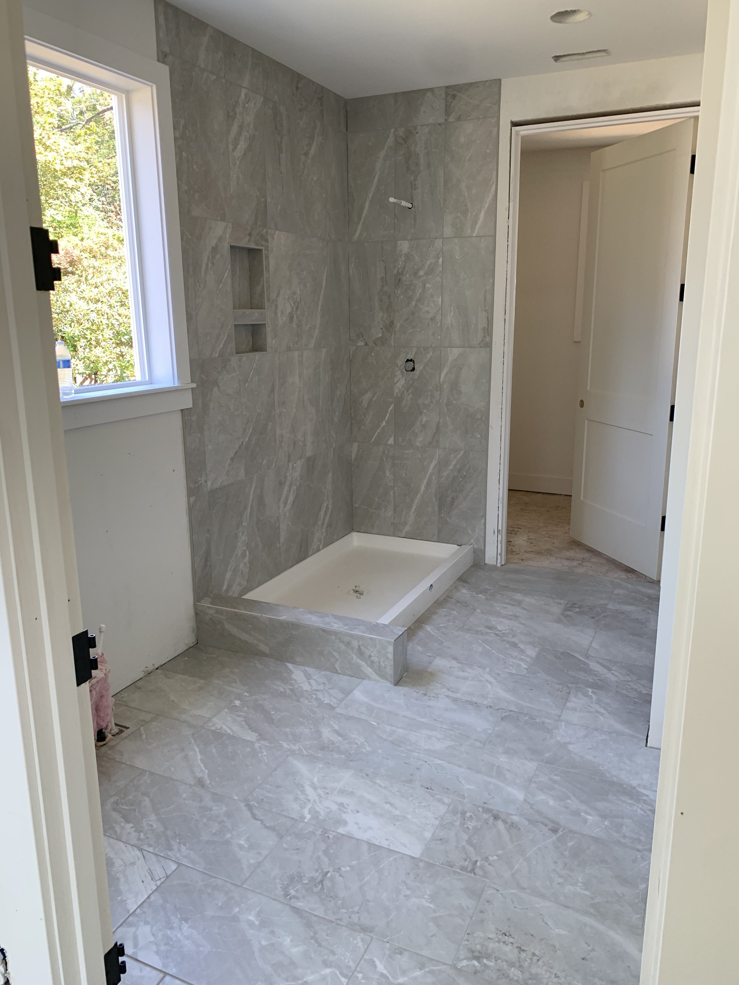 Bathroom under construction with marble tile walls and floors, an empty shower base, and a window showing outside greenery.