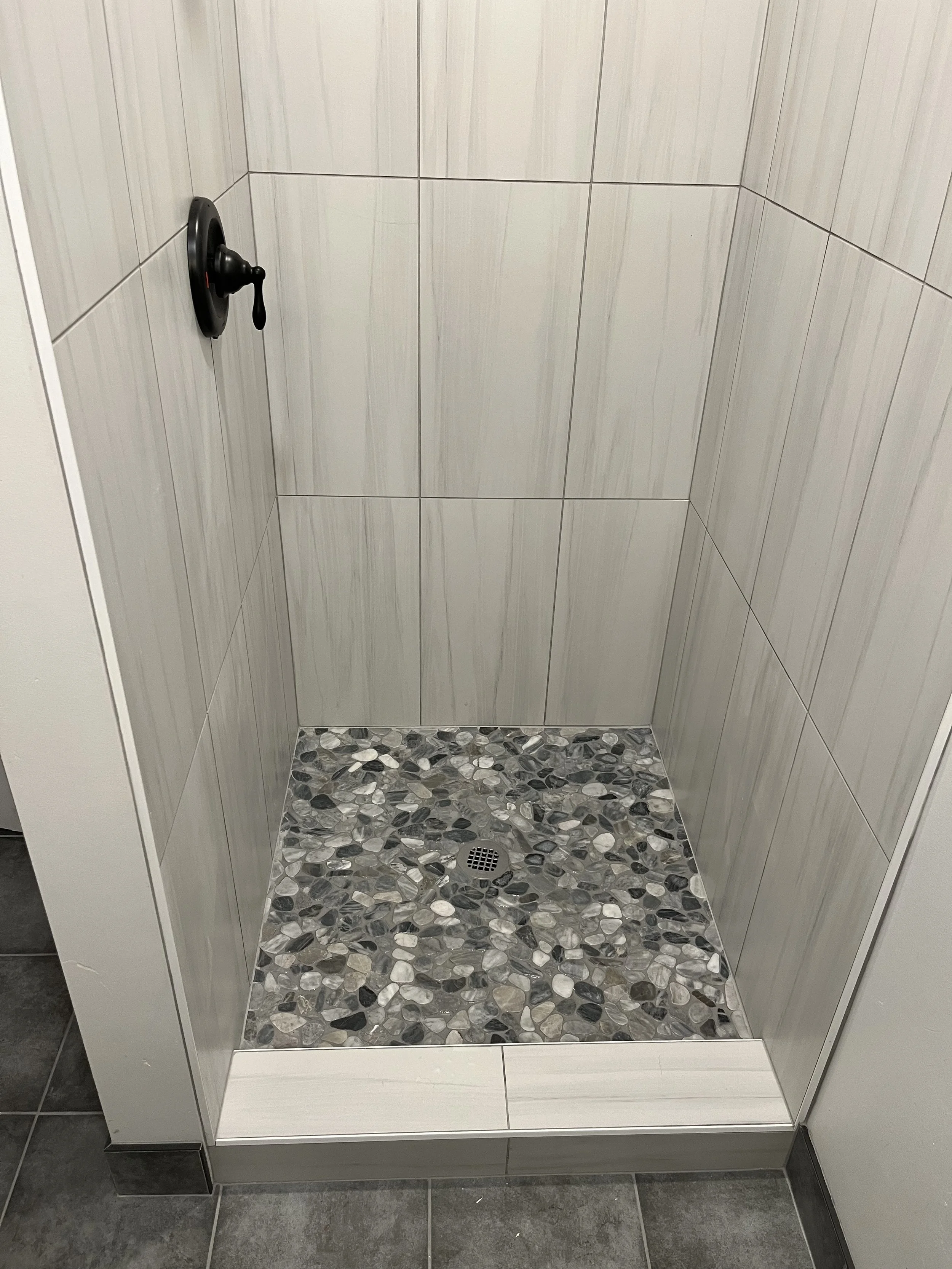Empty shower stall with light-colored tiled walls, mosaic-style pebble floor, and a drain in the center.