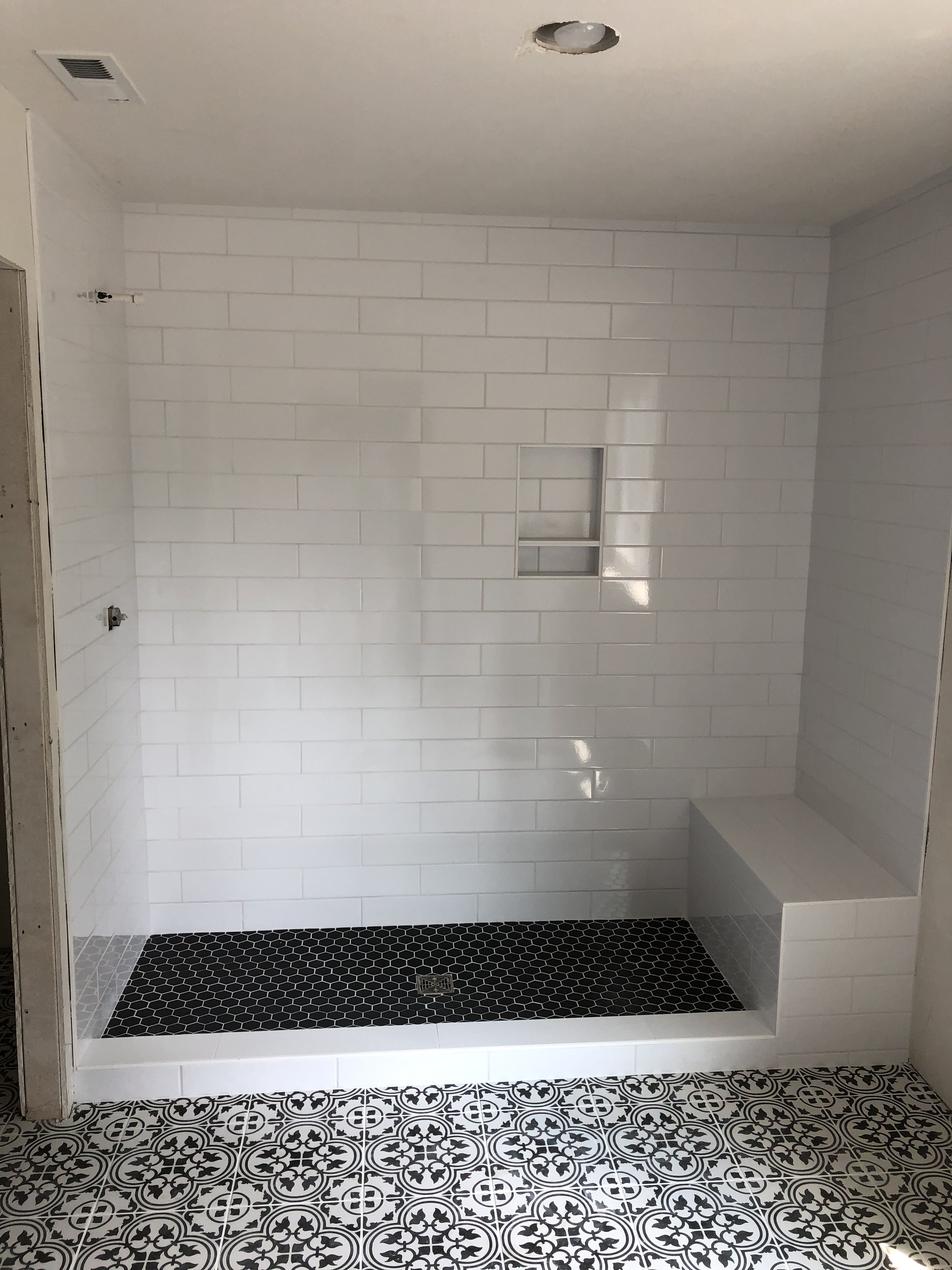 A bathroom with white subway tile walls, a small recessed shelf, and a shower floor with black hexagonal tiles, along with patterned black and white floor tiles outside the shower.