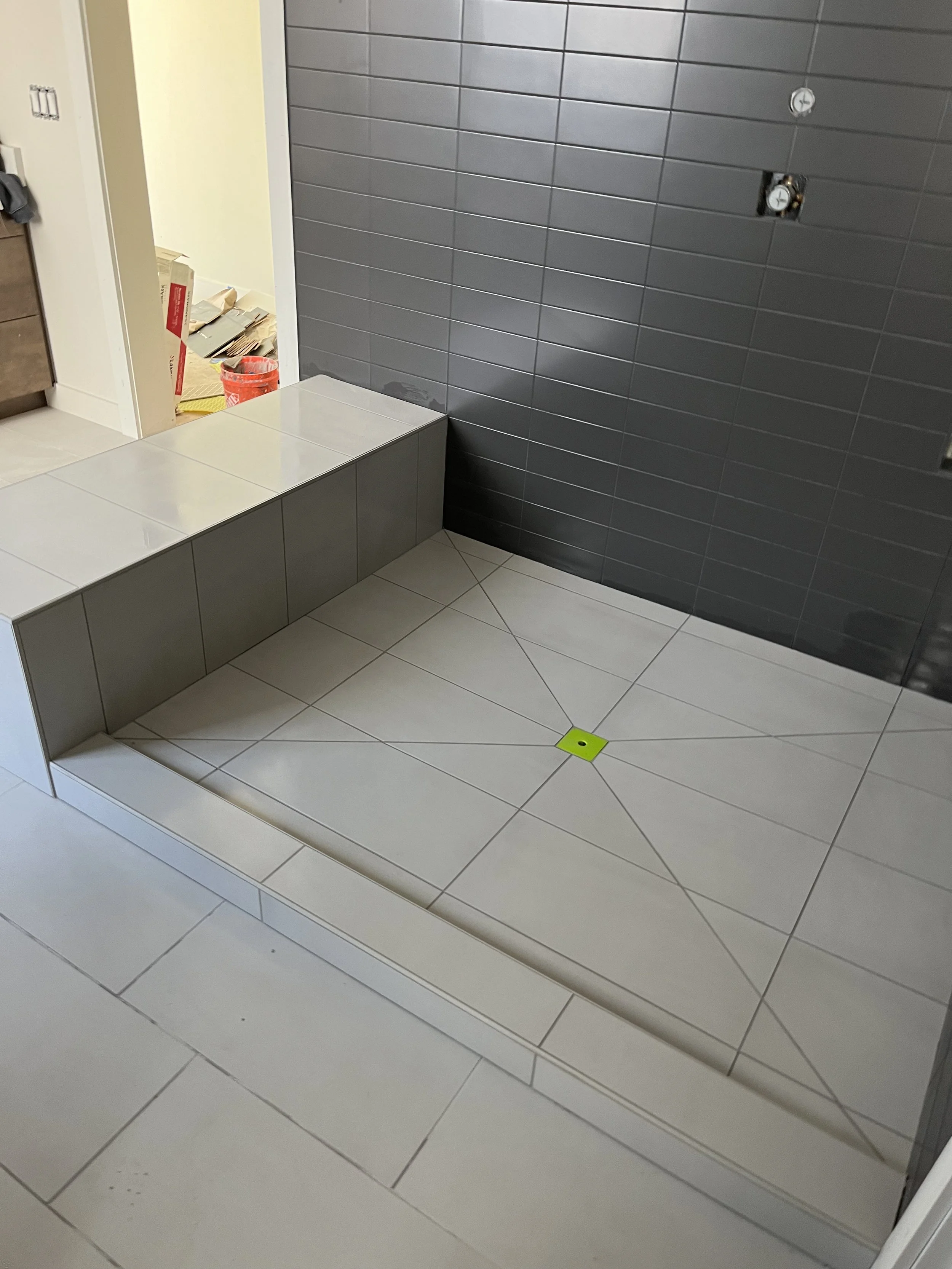 Interior of a shower area with gray tiled walls and a white tiled floor, featuring a bench with gray tiles and a yellow drain cover on the floor.