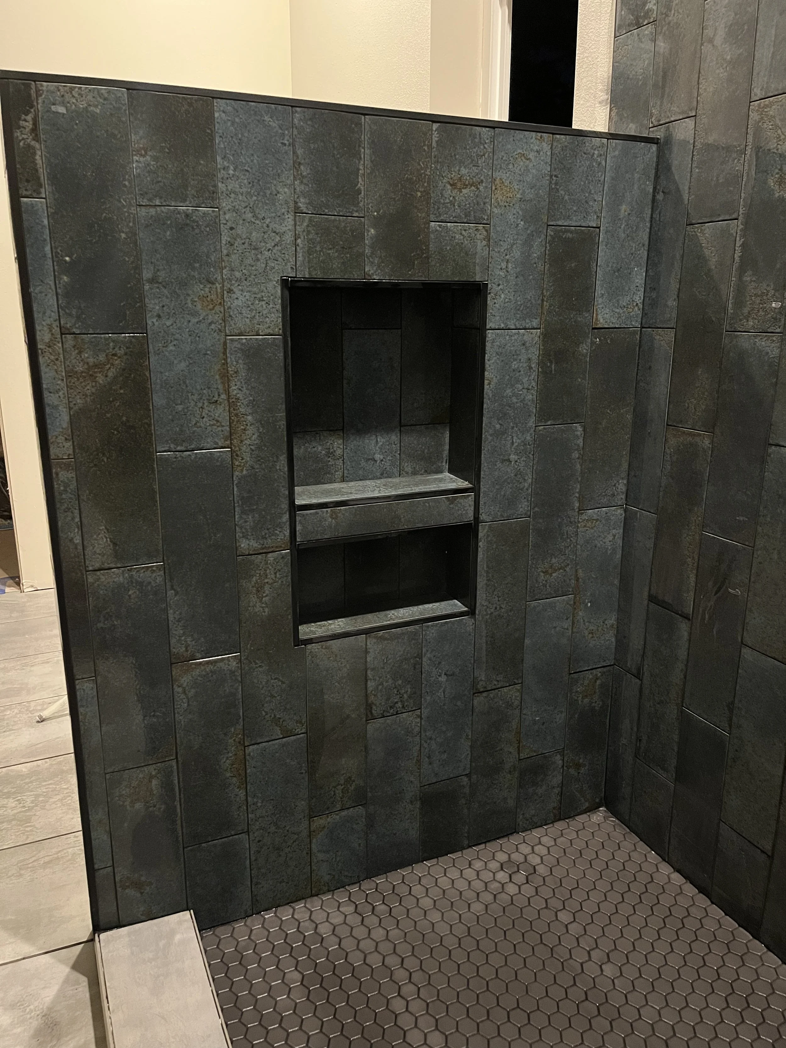 A tiled bathroom shower with dark gray textured tiles on the walls, a small built-in shelf, and light gray hexagonal tiles on the floor.