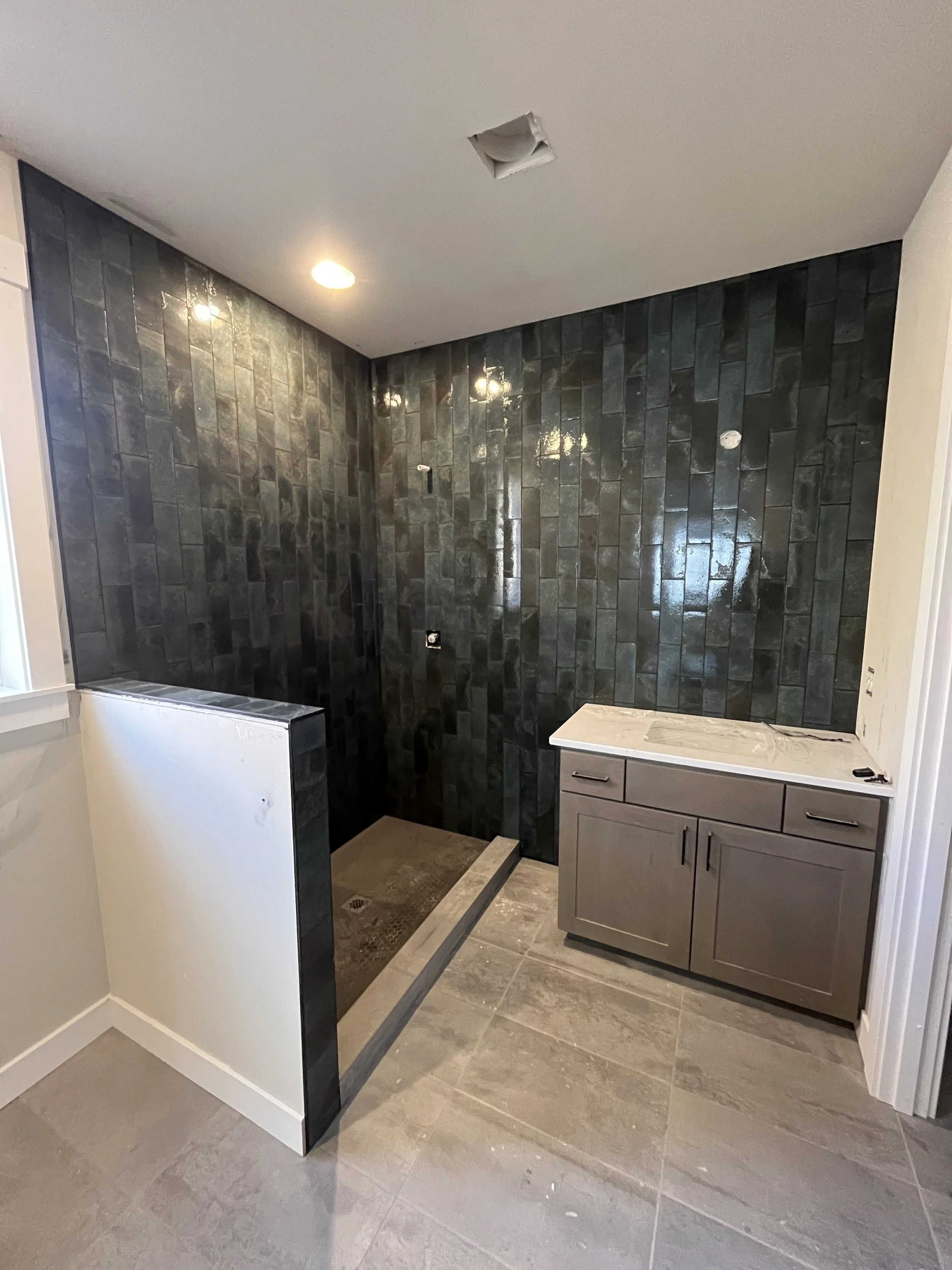 A partially constructed bathroom with a black tiled wall, a sink vanity, and a ceiling with a hole for a light fixture. The floor has large beige tiles.