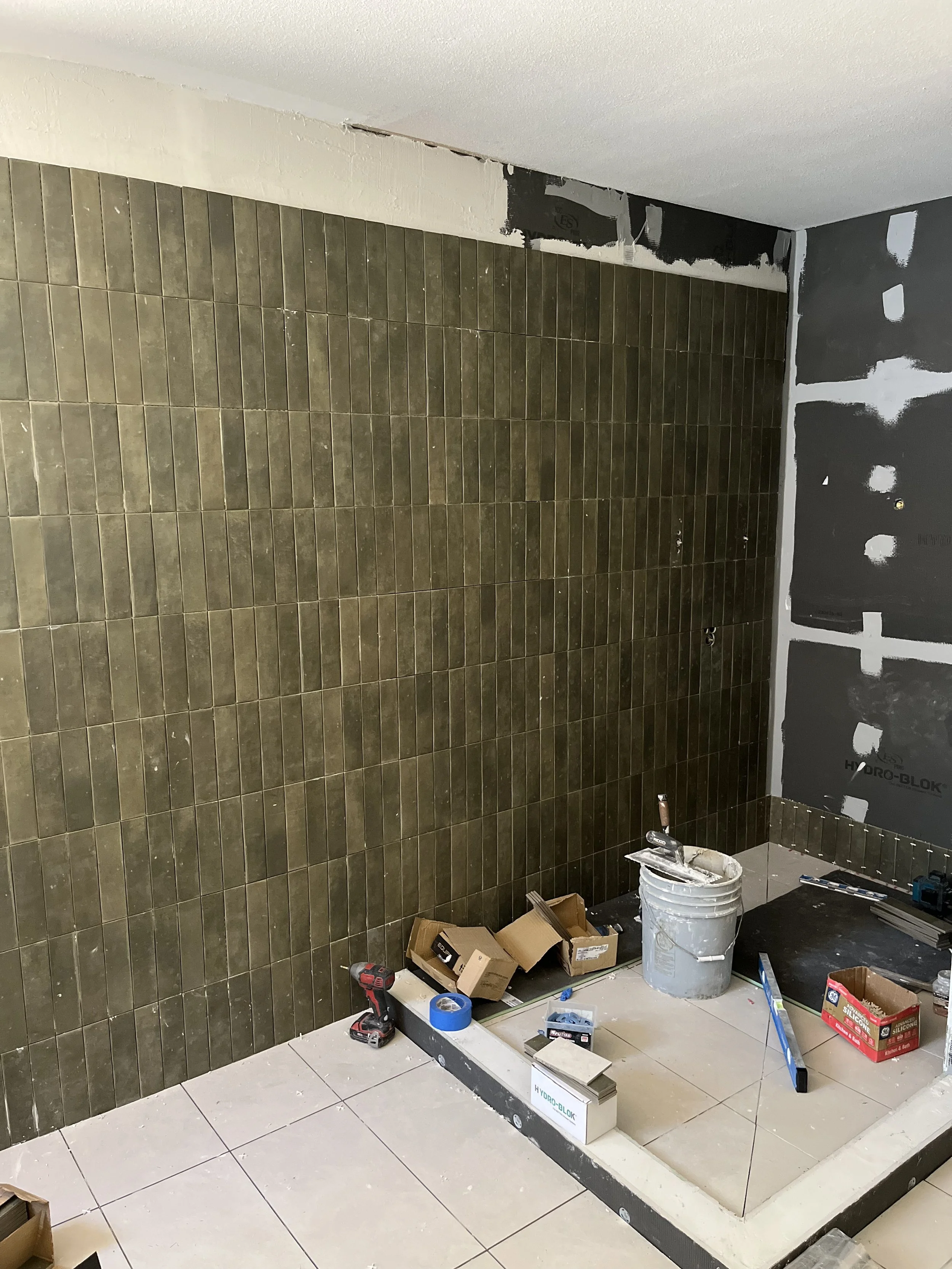 Bathroom under renovation with tiled wall, construction tools, and materials scattered on the floor.