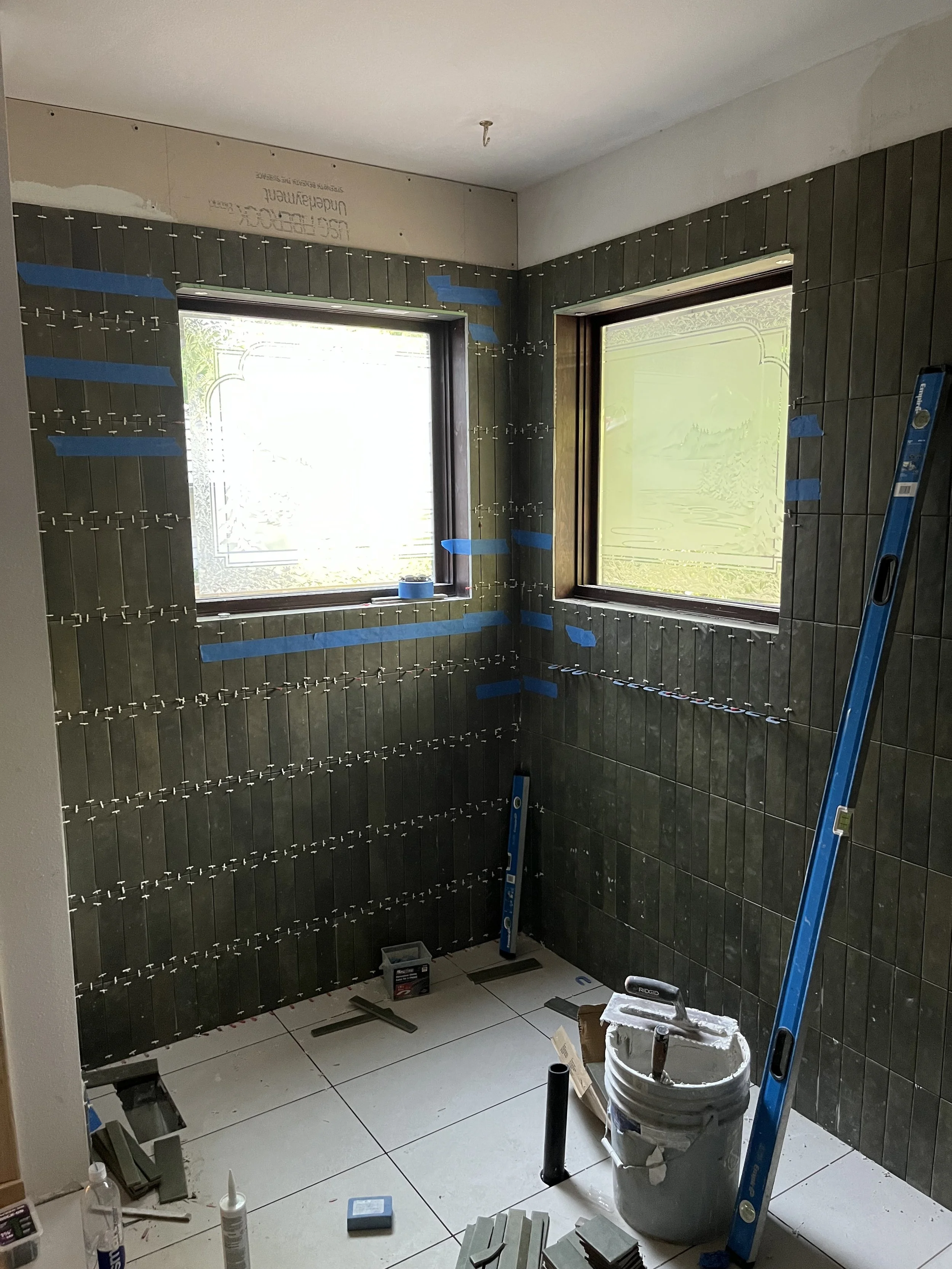 Bathroom wall under construction with dark tiles, blue painter's tape, and clear glass window with a landscape design. Tools and supplies such as a bucket, level, and adhesive materials are on the floor.