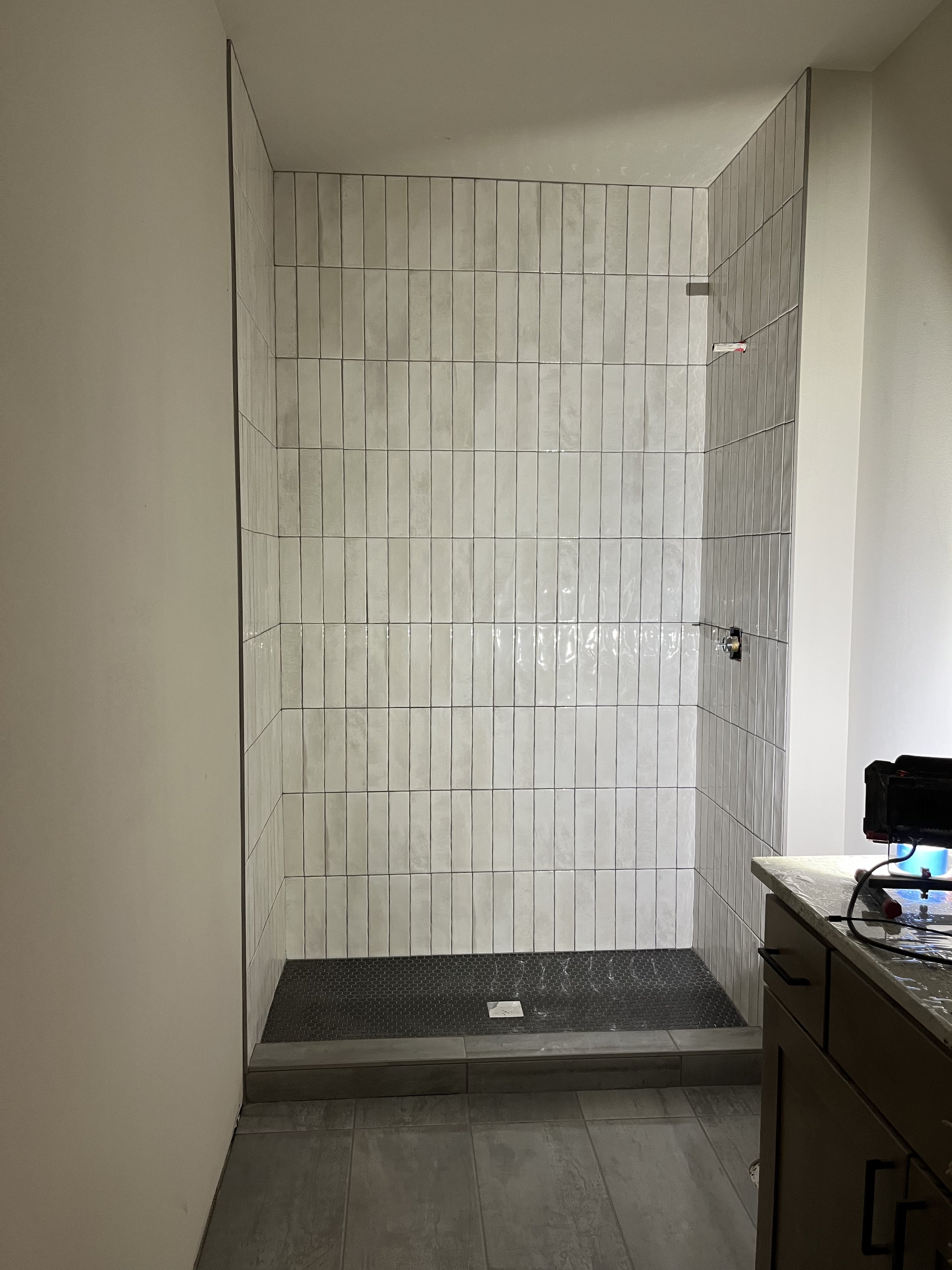 Bathroom shower area with tiled walls, a black rubber mat on the floor, and a small countertop with items on it.