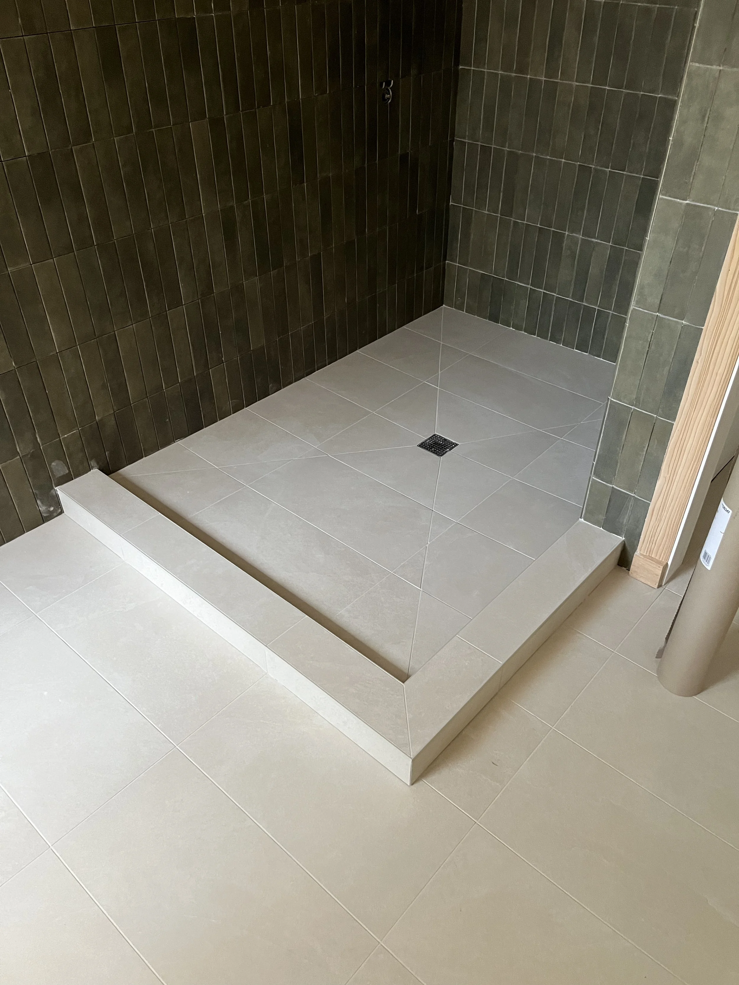 A walk-in shower with beige floor tiles, dark green wall tiles, and a small square drain on the floor. A raised ledge surrounds part of the shower area.