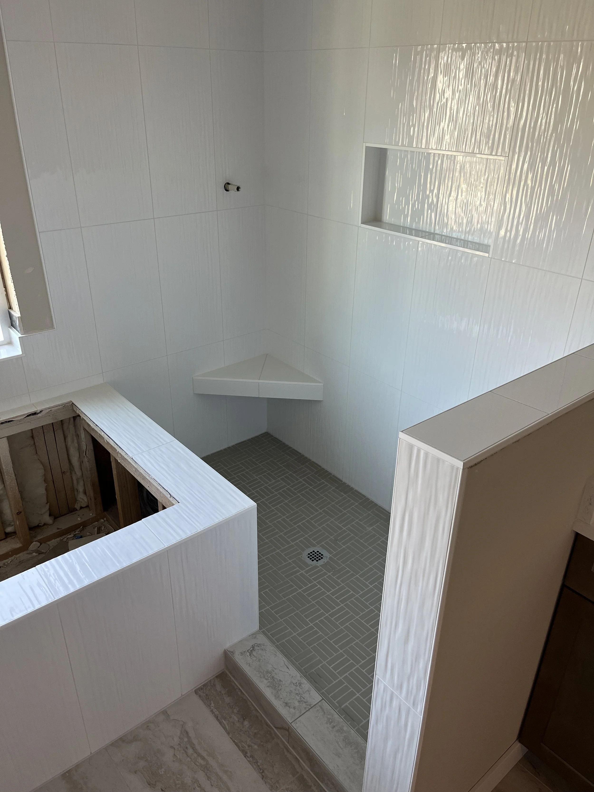Bathroom under construction with white tiled walls, a small shower area with gray tiles, and an open wall under renovation.