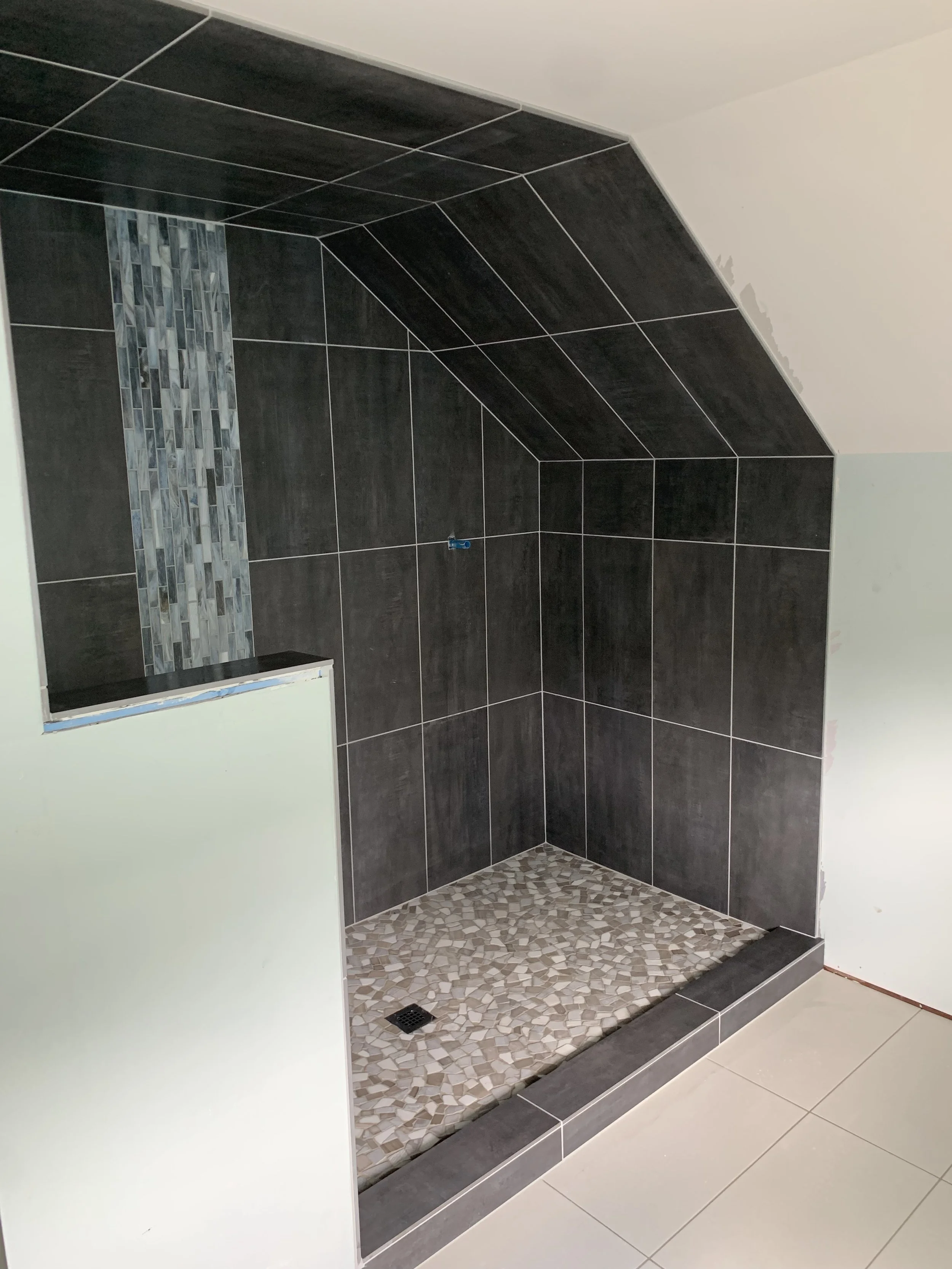 Bathroom or shower area with dark gray tiles on the wall and a decorative strip of small gray and white tiles. The shower floor has small pebble-like tiles, and there is a drain at the center. Part of a white wall and tiled floor are visible outside 