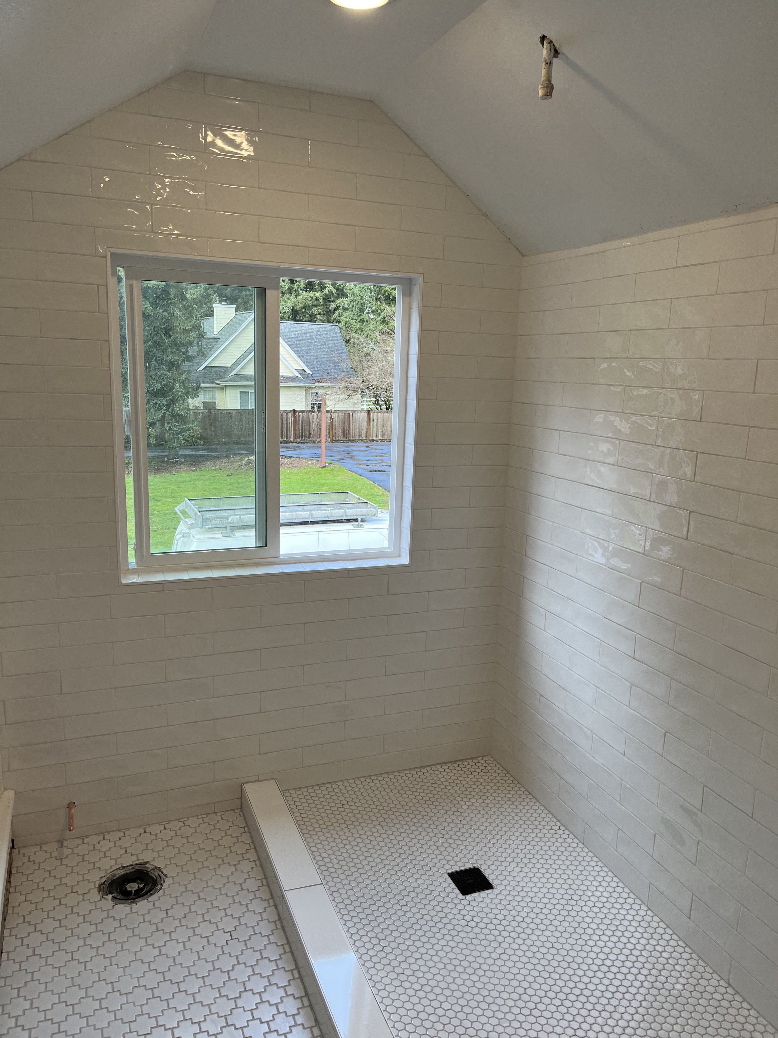 Bathroom with white subway tile walls, window overlooking a yard, and a shower area with hexagonal and penny tiles, with plumbing fixtures and a drainage grate.