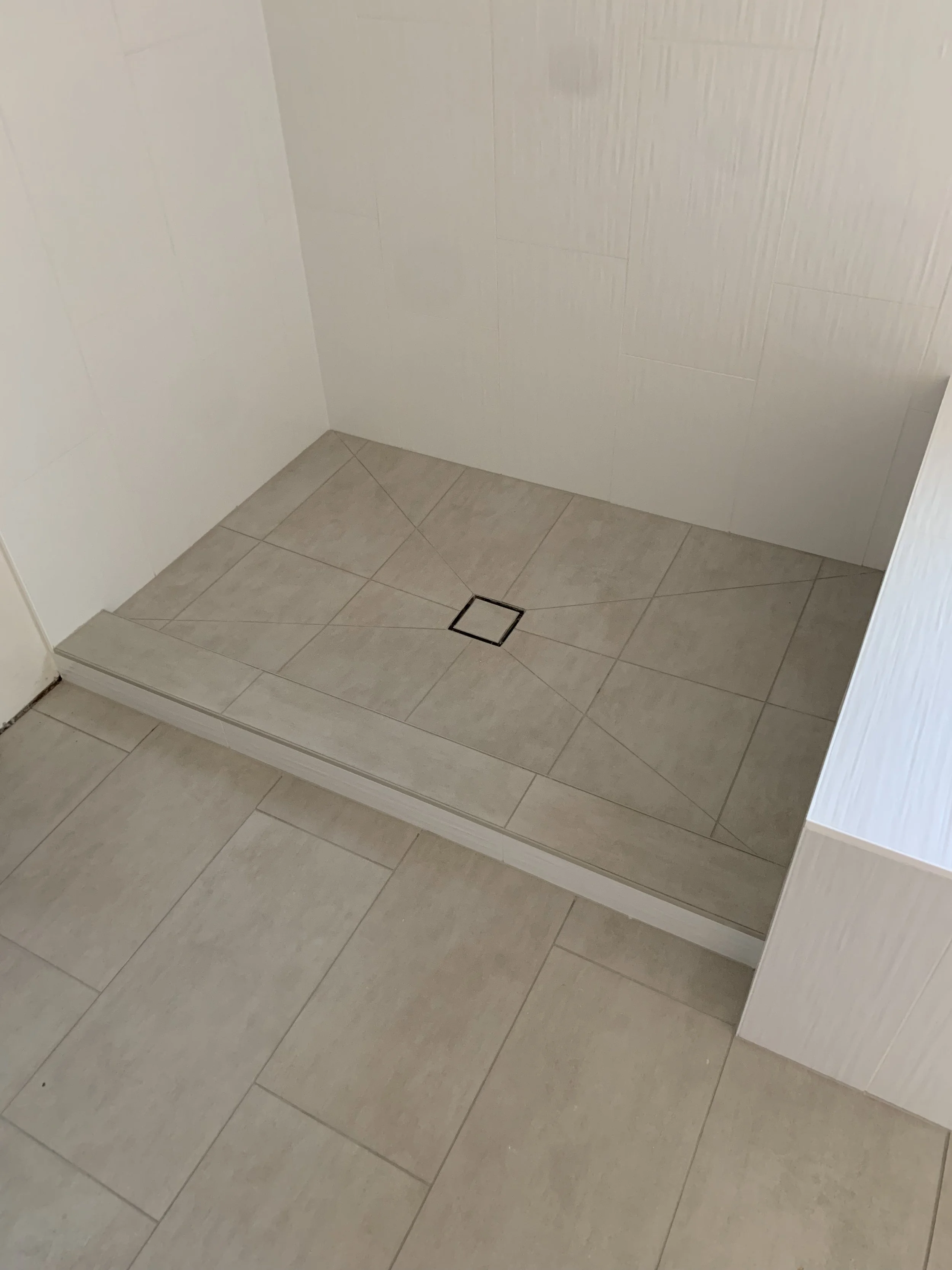 A beige tiled bathroom floor with a drainage grate and a small step up to a slightly different tile level.
