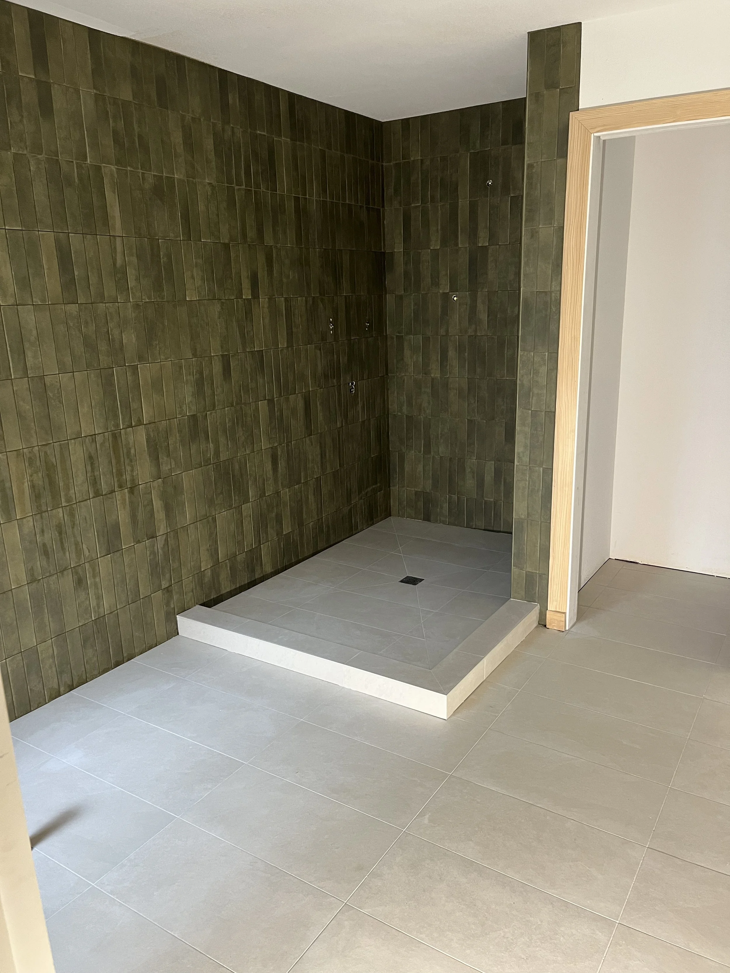Empty shower stall with dark green rectangular tiles and a small drain at the center. The shower area is slightly elevated with a white border, adjacent to a beige tiled bathroom floor and an opening to a white wall with a wooden door frame.