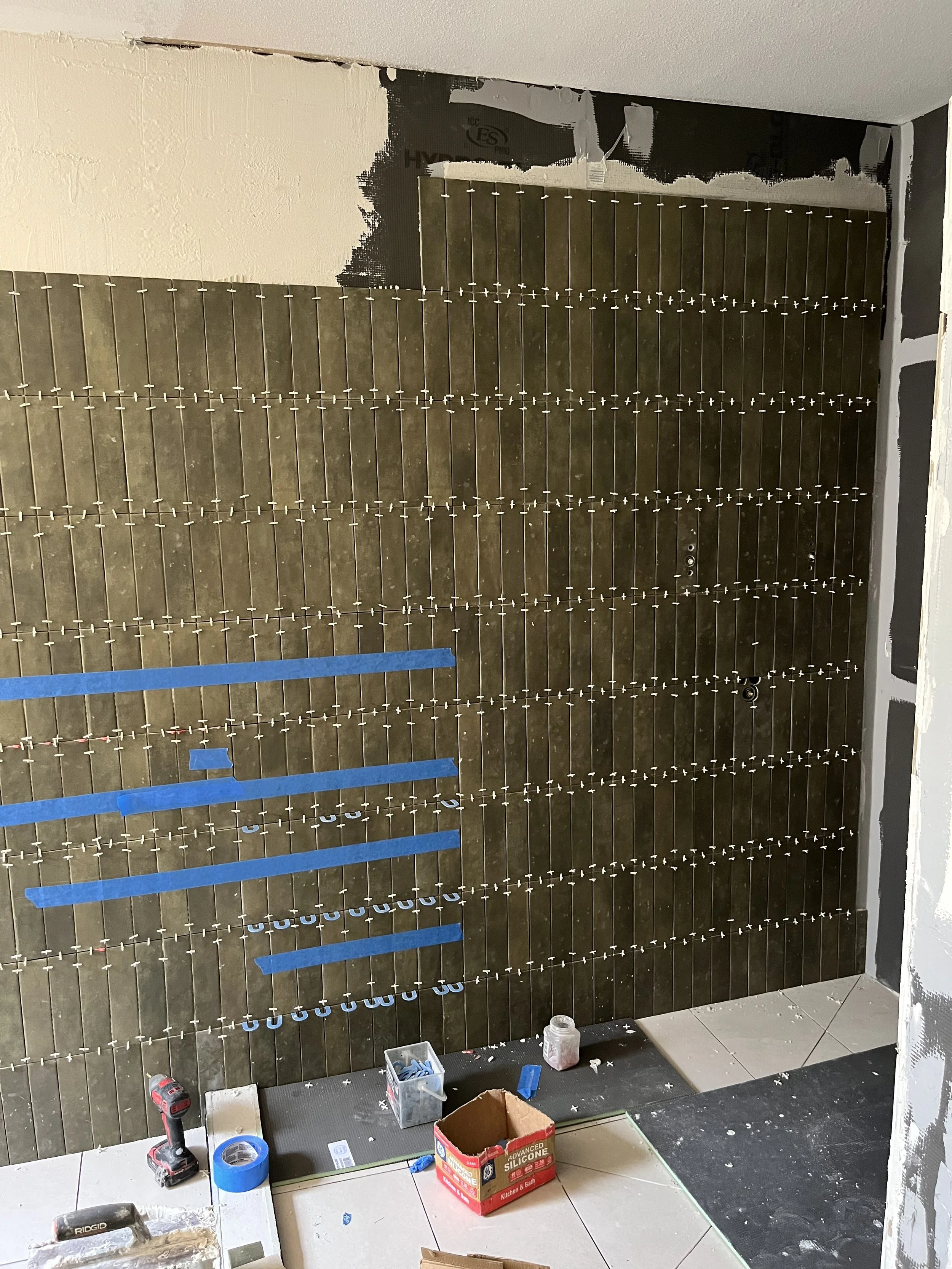 A wall during renovation, with tiles being installed, tools and materials scattered on the floor.