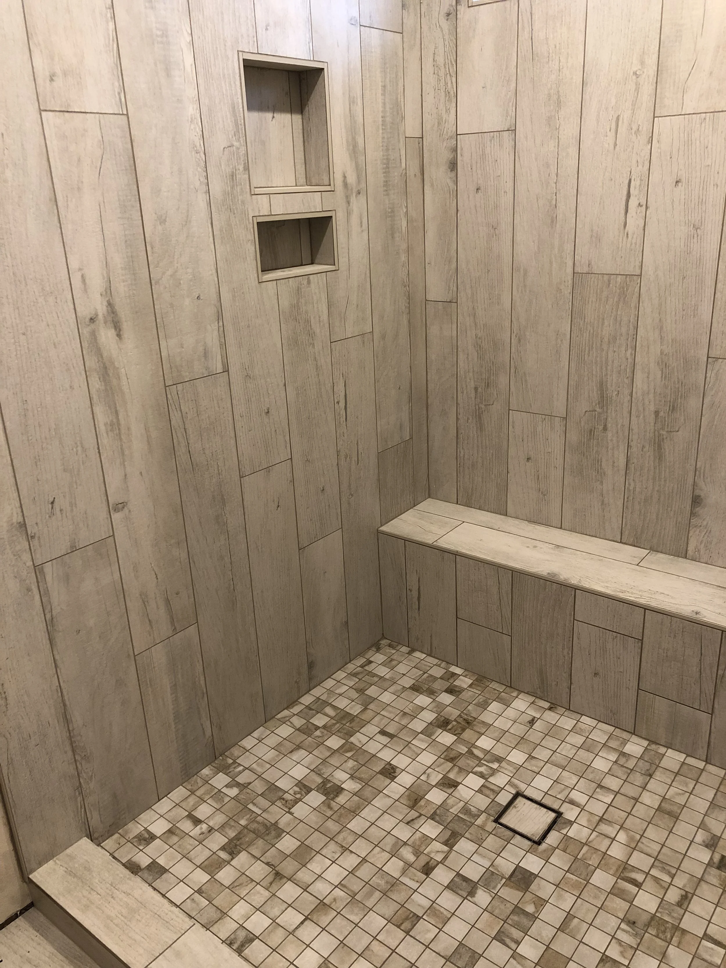 A bathroom shower with wall tiles that resemble light wood and a small mosaic tile floor. There are two recessed shelves built into the wall, and a square drain on the floor.