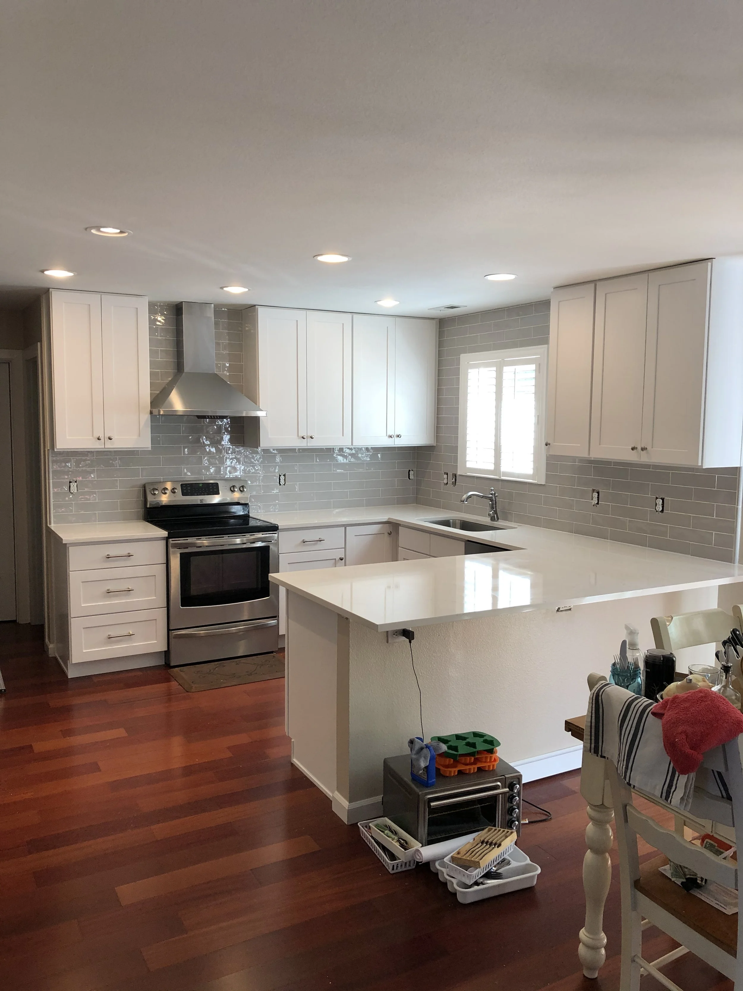 A kitchen with white cabinets, stainless steel oven, gray subway tile backsplash, and a window with plantation shutters. There is a white kitchen island with various items and a dining table with chairs, some items, and a red towel on one chair. The floor is dark wood.