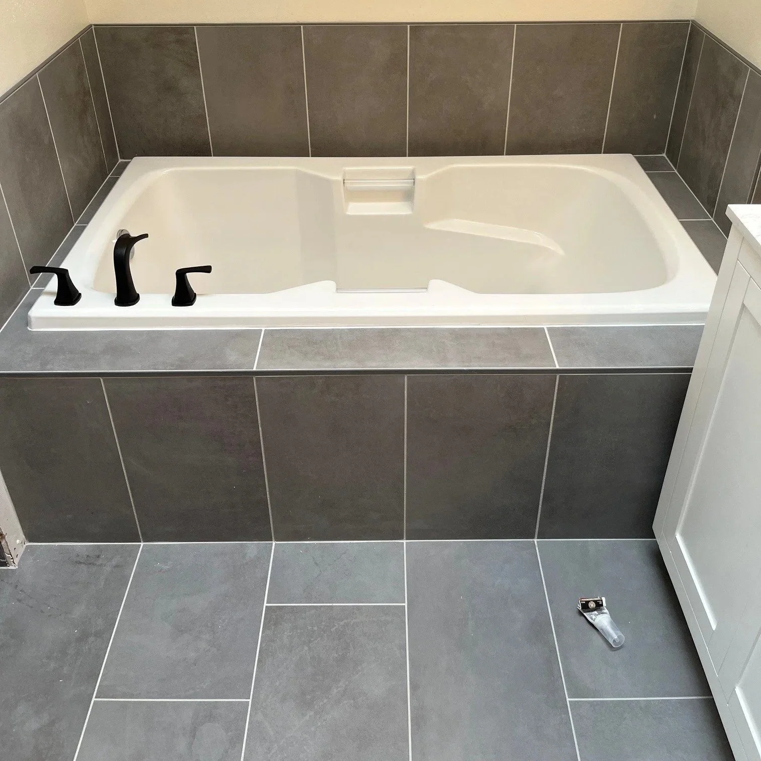 A bathroom with a white bathtub and dark gray tiled walls and floor. The bathtub has black faucet fixtures and a ledge at the back. There is a tube of caulk on the floor in front of the tub.