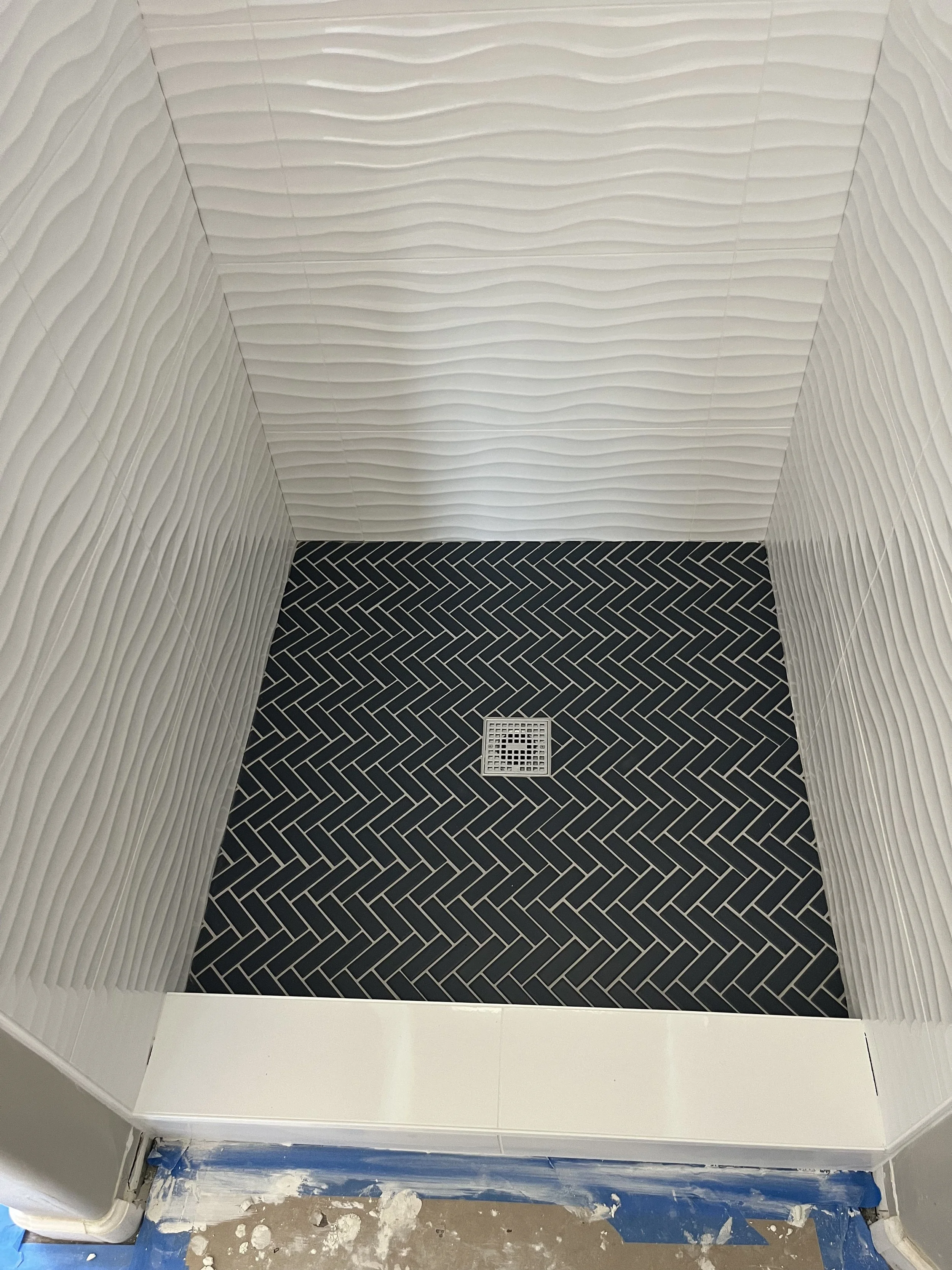 Interior of a bathroom shower area with white textured tiled walls and black herringbone patterned floor tiles, with a drain in the center.