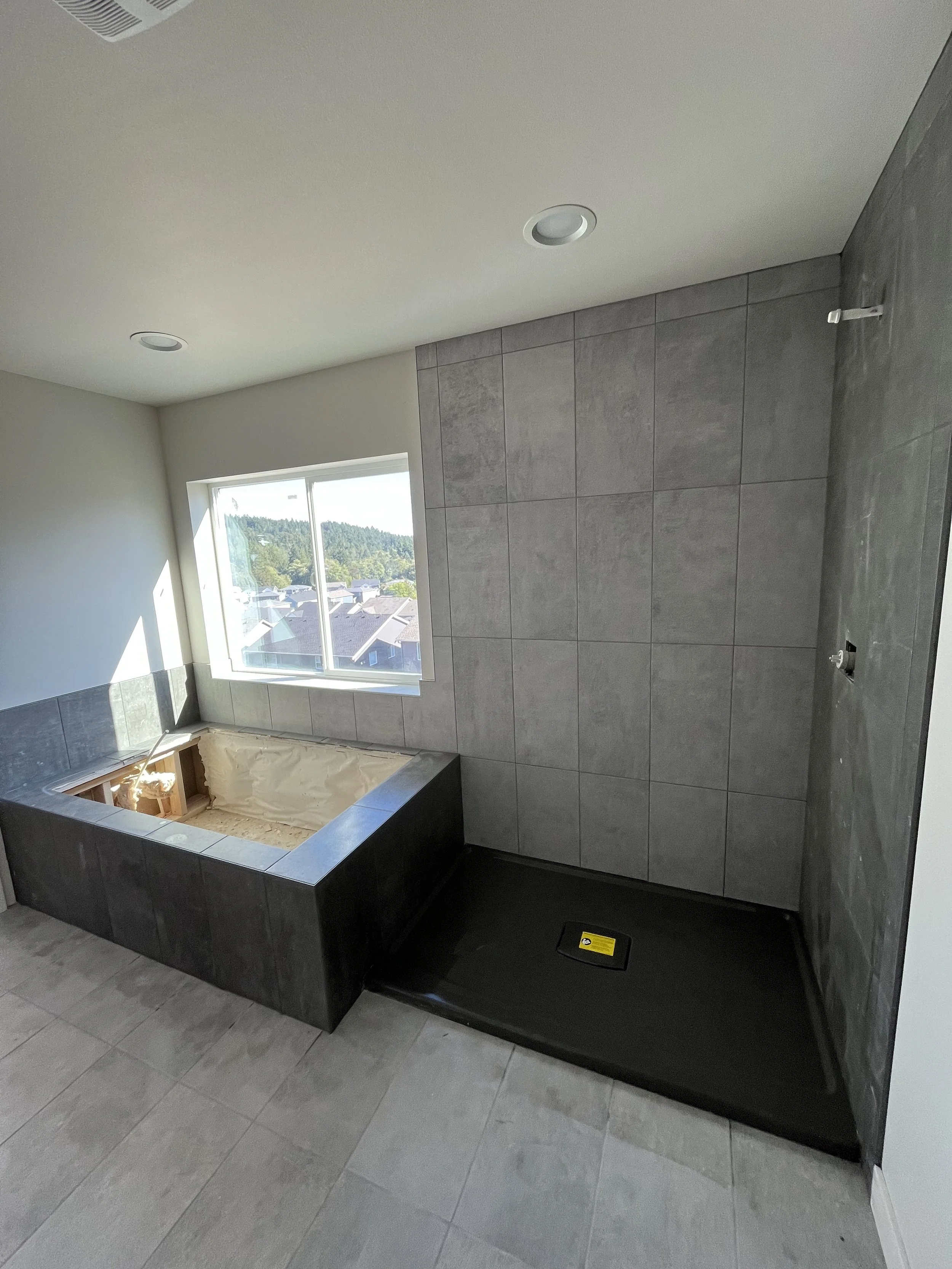 Bathroom under construction with a large window, tiled walls, and a bathtub with a black exterior, without fixtures installed.