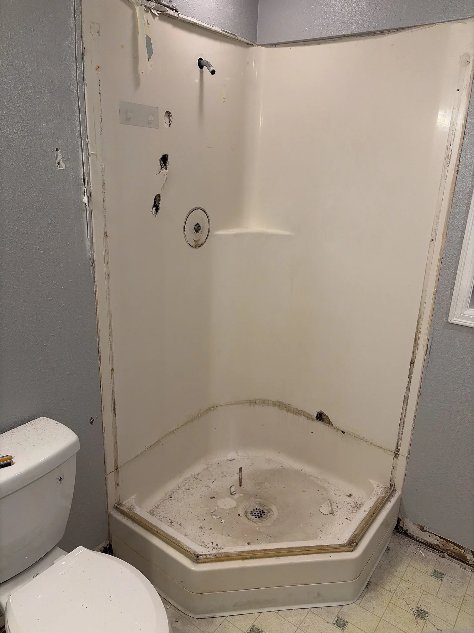 Old, dirty shower with missing fixtures and black mold, next to a toilet with a closed lid, in a bathroom with yellow tiled floor.