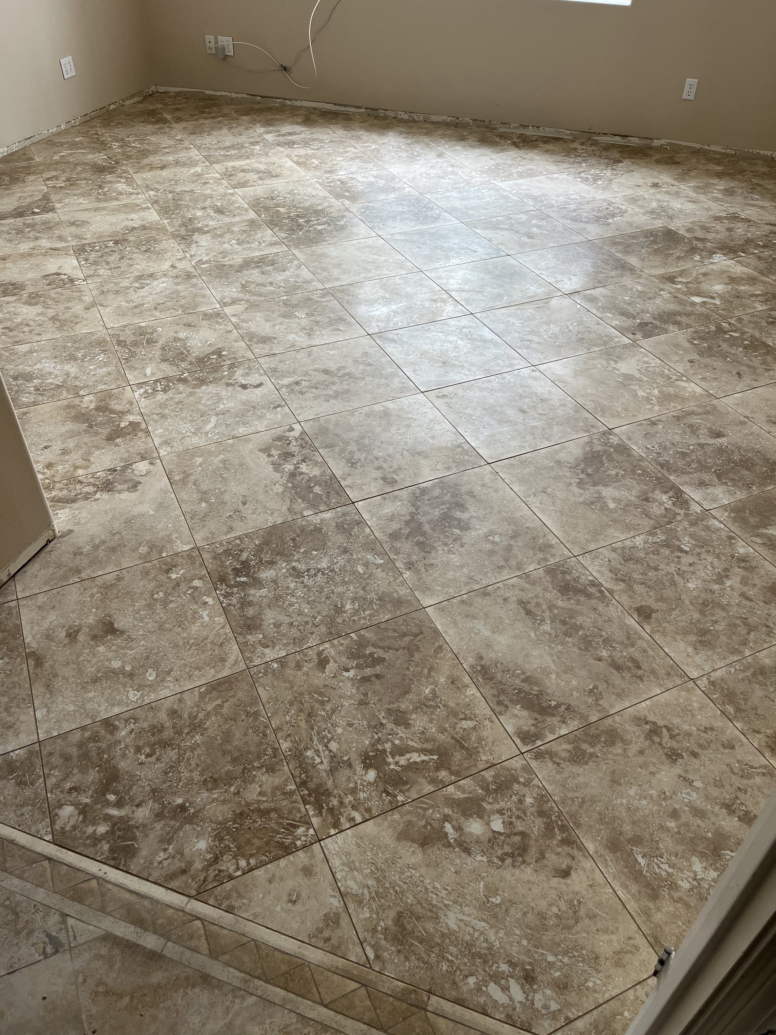 A room with beige walls and electrical outlets. The floor has large, tan and gray marble tiles. There is some unfinished construction along the base of the walls and an extension cord or wire hanging from the wall.