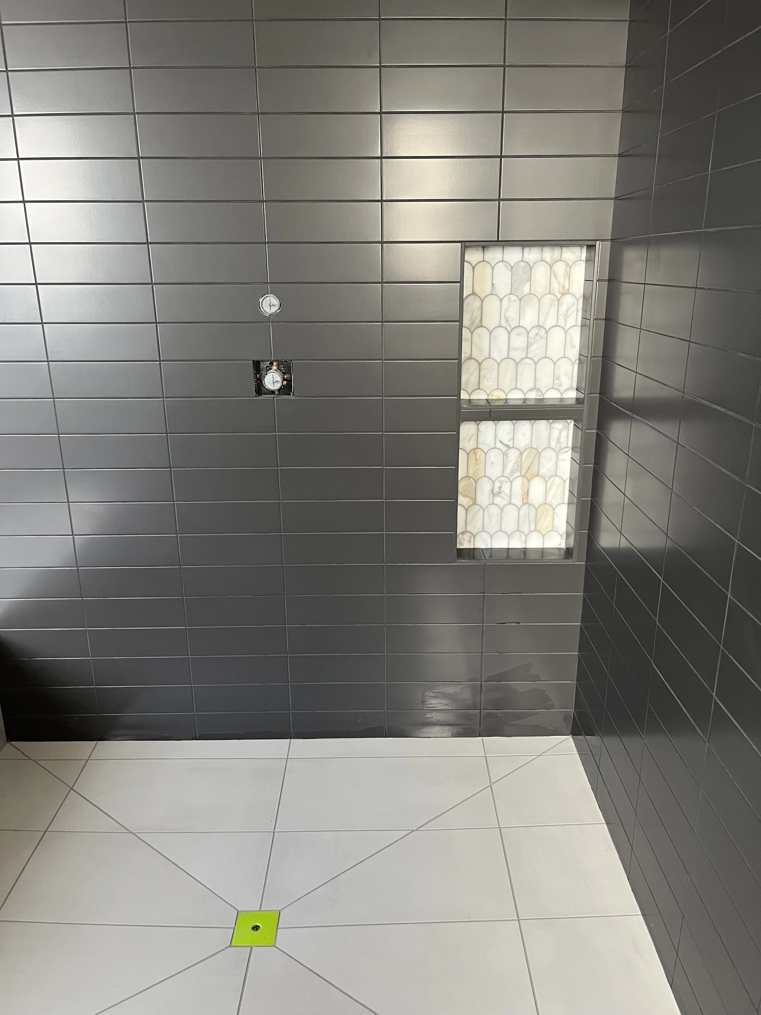A tiled bathroom with gray tiles on the walls. There is a small window with frosted glass blocks on the right side. The floor has large white tiles with a small bright green square drain cover near the center.
