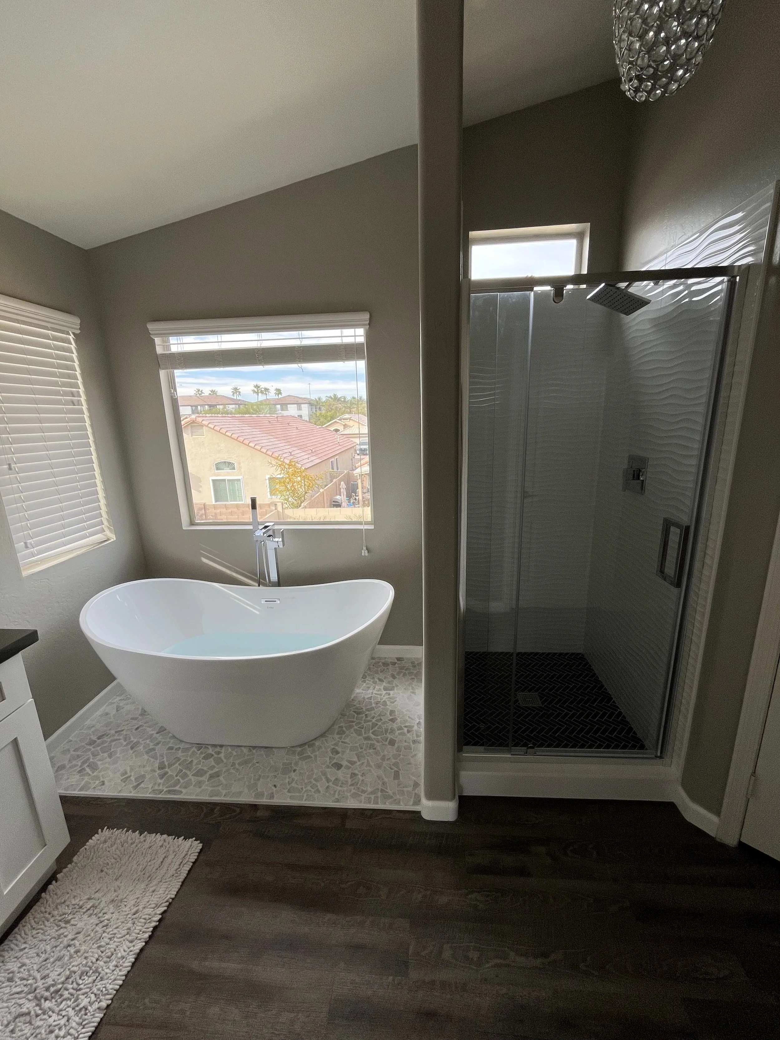 Modern bathroom with freestanding bathtub, window showing a neighborhood view, and a glass enclosed shower with textured wall tiles and a rainfall showerhead.