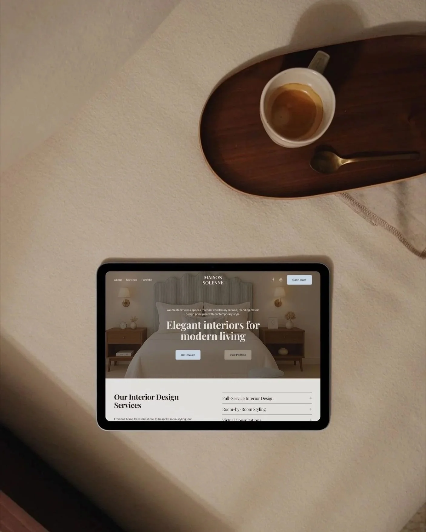 Tablet displaying a website for interior design services, placed on a wooden tray on a beige wall shelf. A coffee mug with coffee remnants and a spoon are also on the tray.