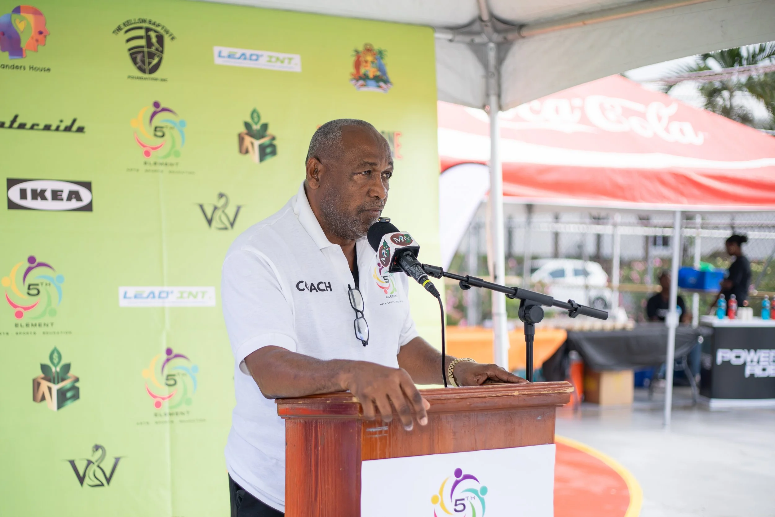 A man in a white shirt labeled 'COACH' standing at a podium, speaking into a microphone at an outdoor event with a green backdrop and various logos behind him.