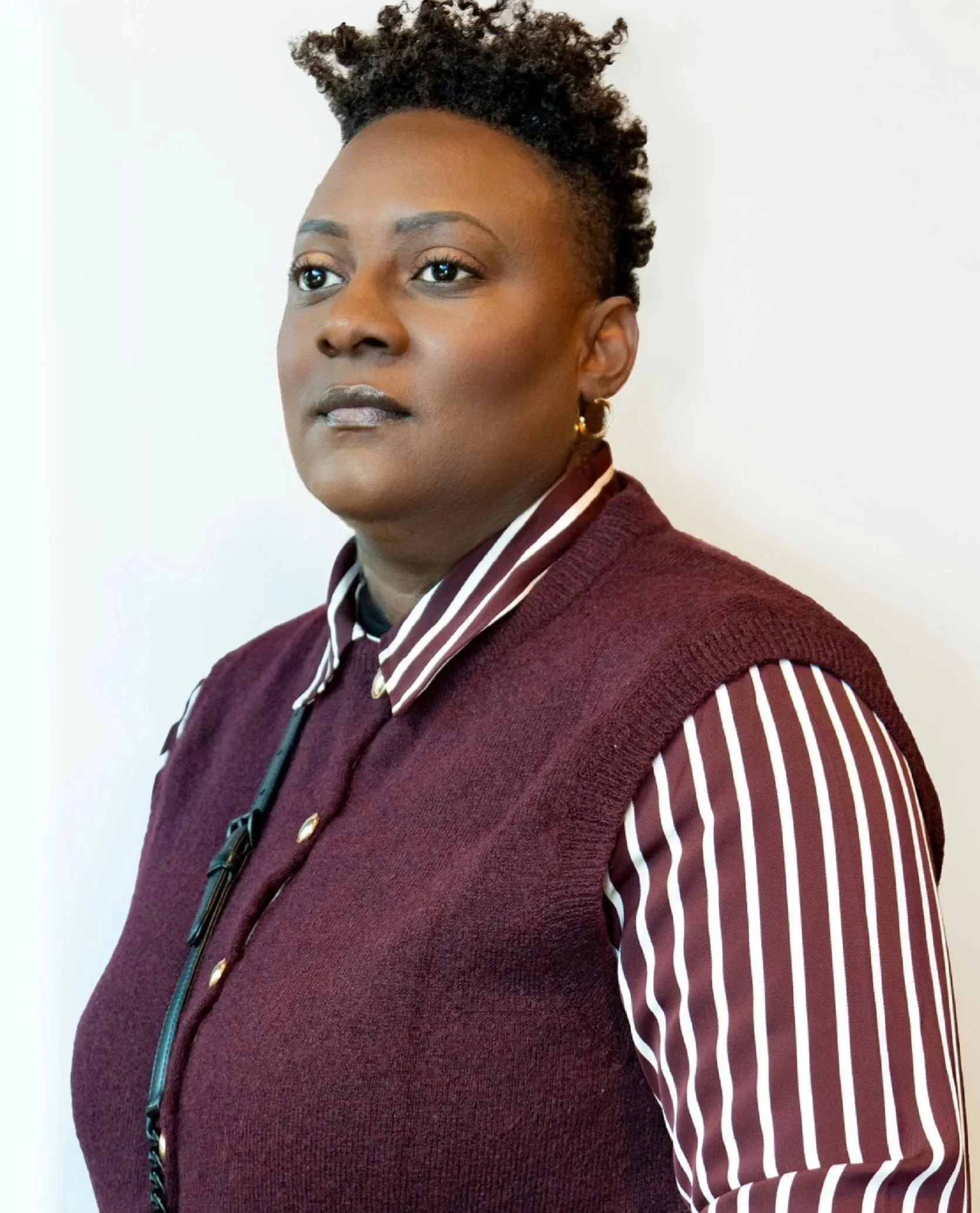 A woman with dark skin, long dreadlocks, wearing a black pinstripe blazer and a pearl necklace, taking a selfie indoors in front of a plain wall.