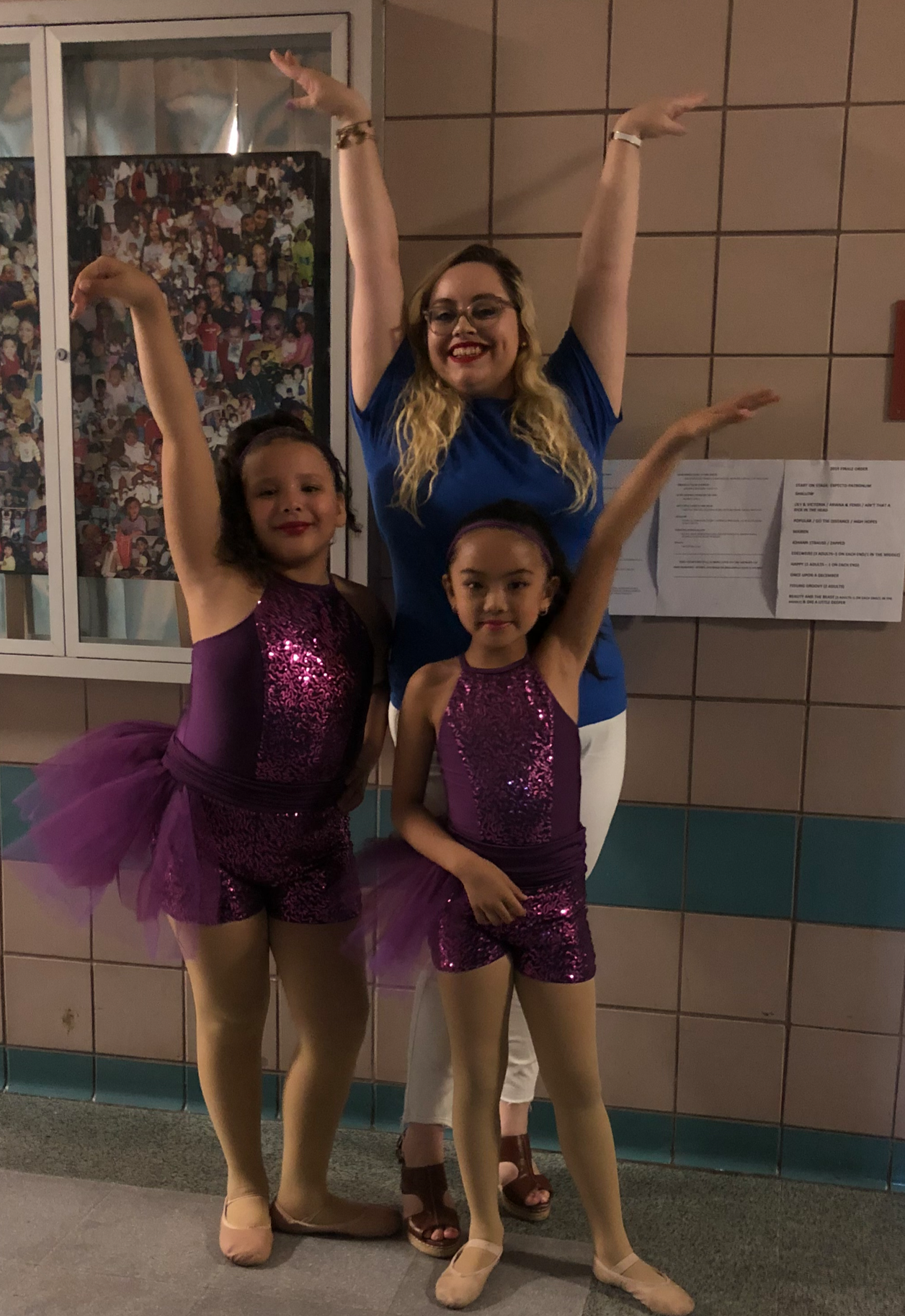 Three women, two young girls dressed in purple sequined ballet costumes with tutus, posing together indoors in front of a tiled wall and a glass cabinet with photographs, smiling with their arms raised.