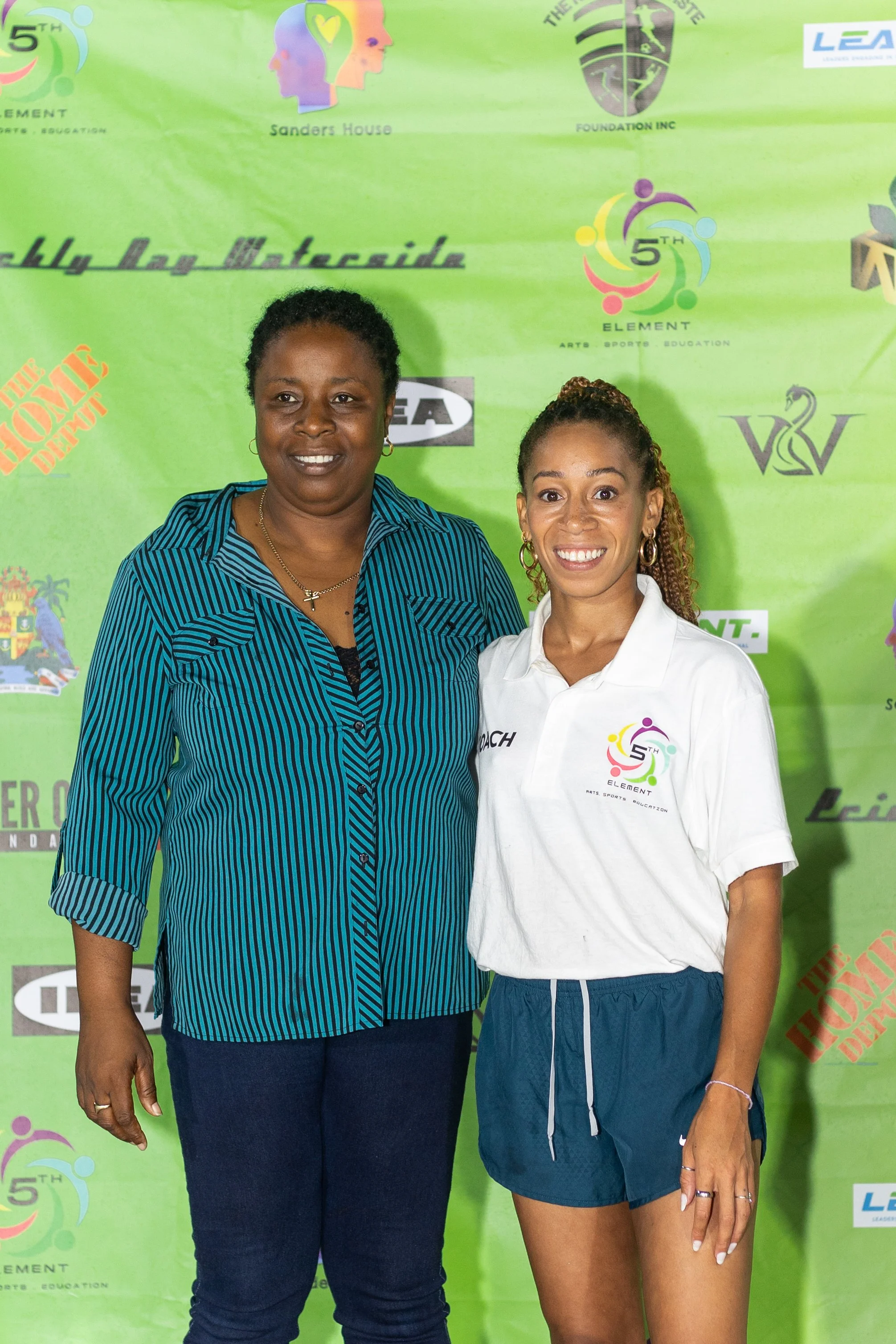 Two women standing in front of a green event banner, smiling. One woman wears a blue striped shirt, the other a white polo with a logo.