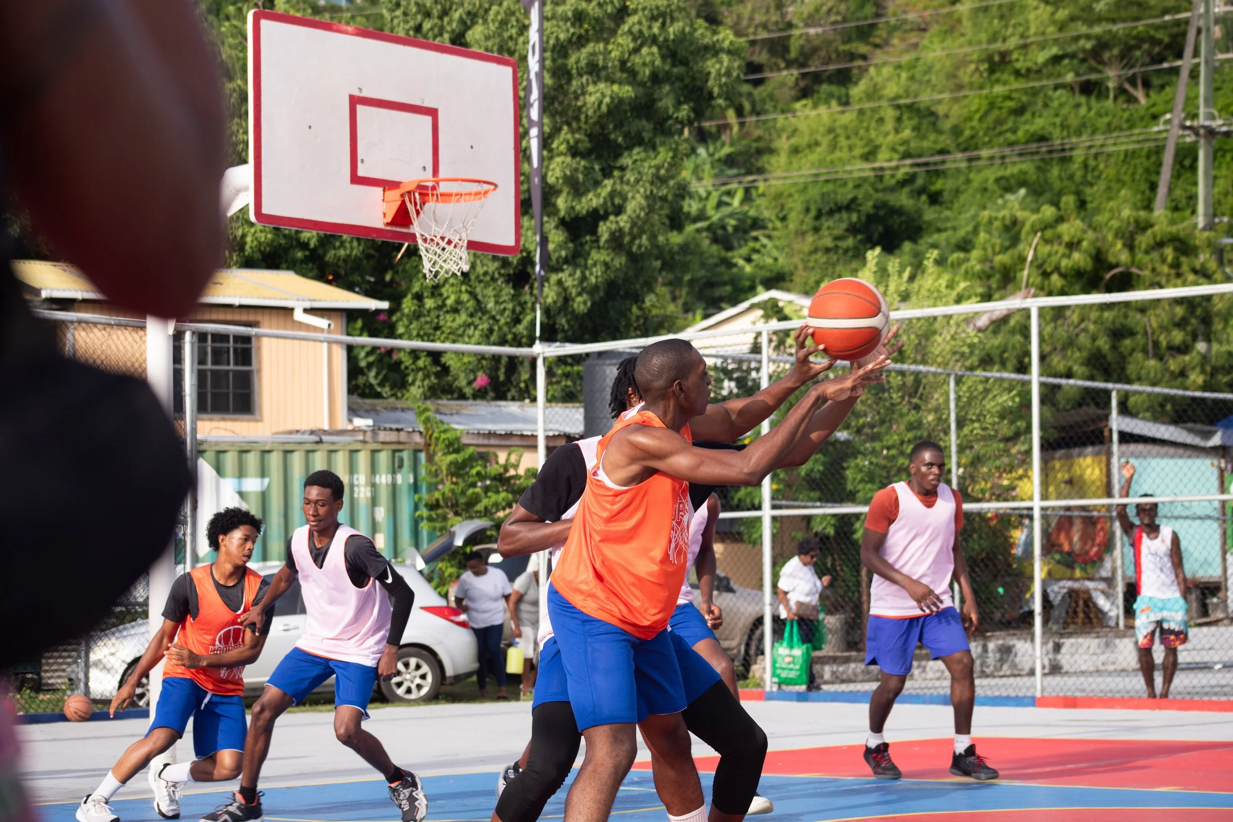 A group of young men playing basketball on an outdoor court, with one player preparing to shoot the ball. Others are watching or guarding, with a backboard and hoop visible, surrounded by greenery and residential buildings.