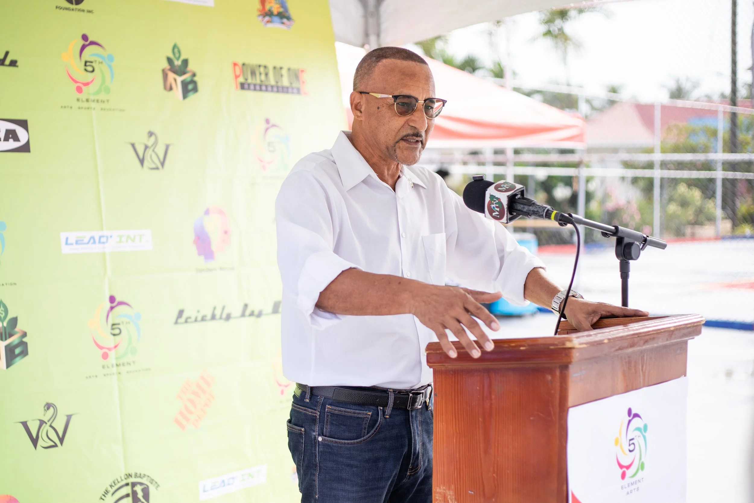 A man in a white shirt and sunglasses speaking at a podium with a microphone during an outdoor event.
