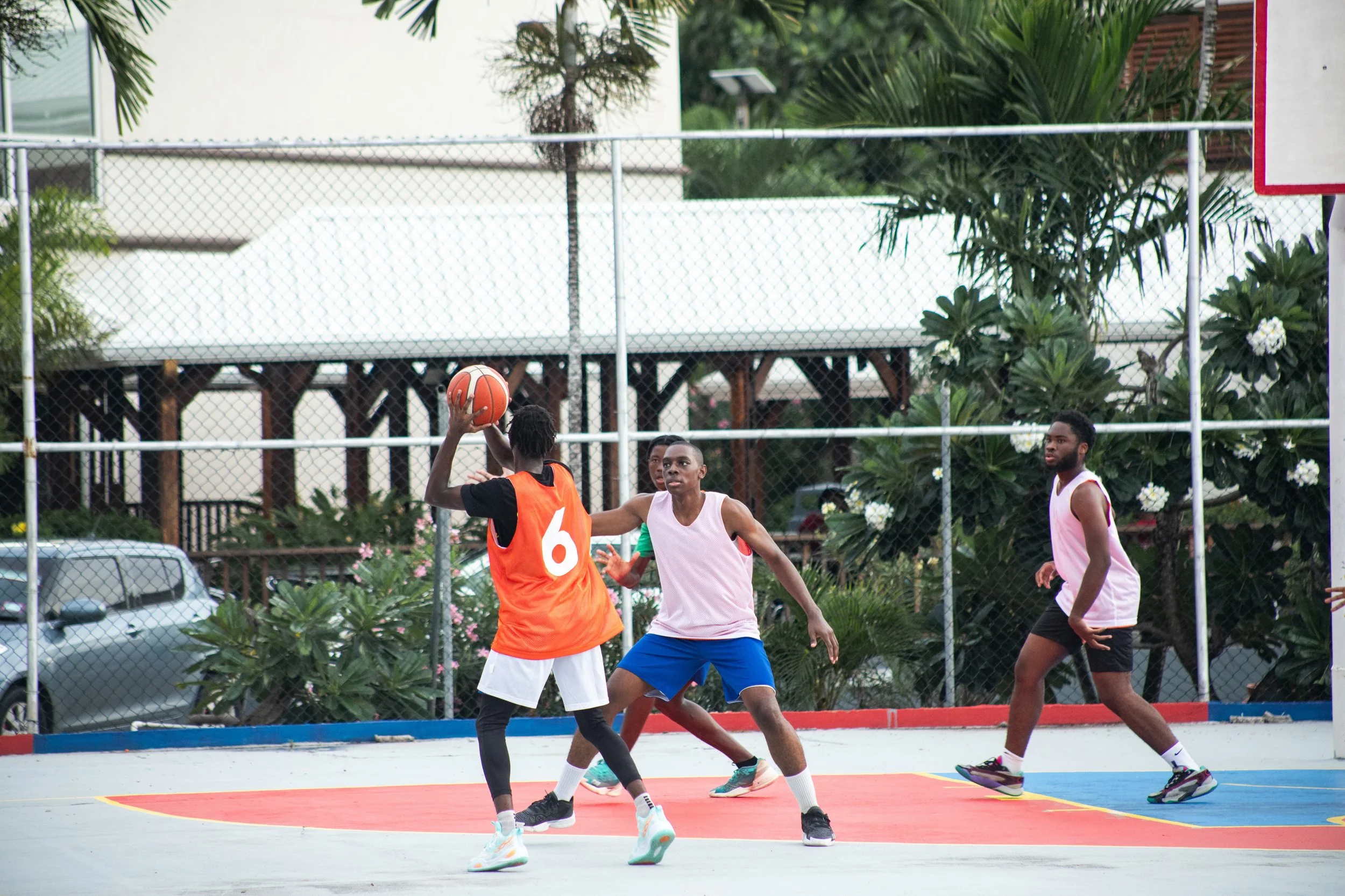 A group of young men playing basketball on an outdoor court surrounded by a chain-link fence, with trees and parked cars in the background.