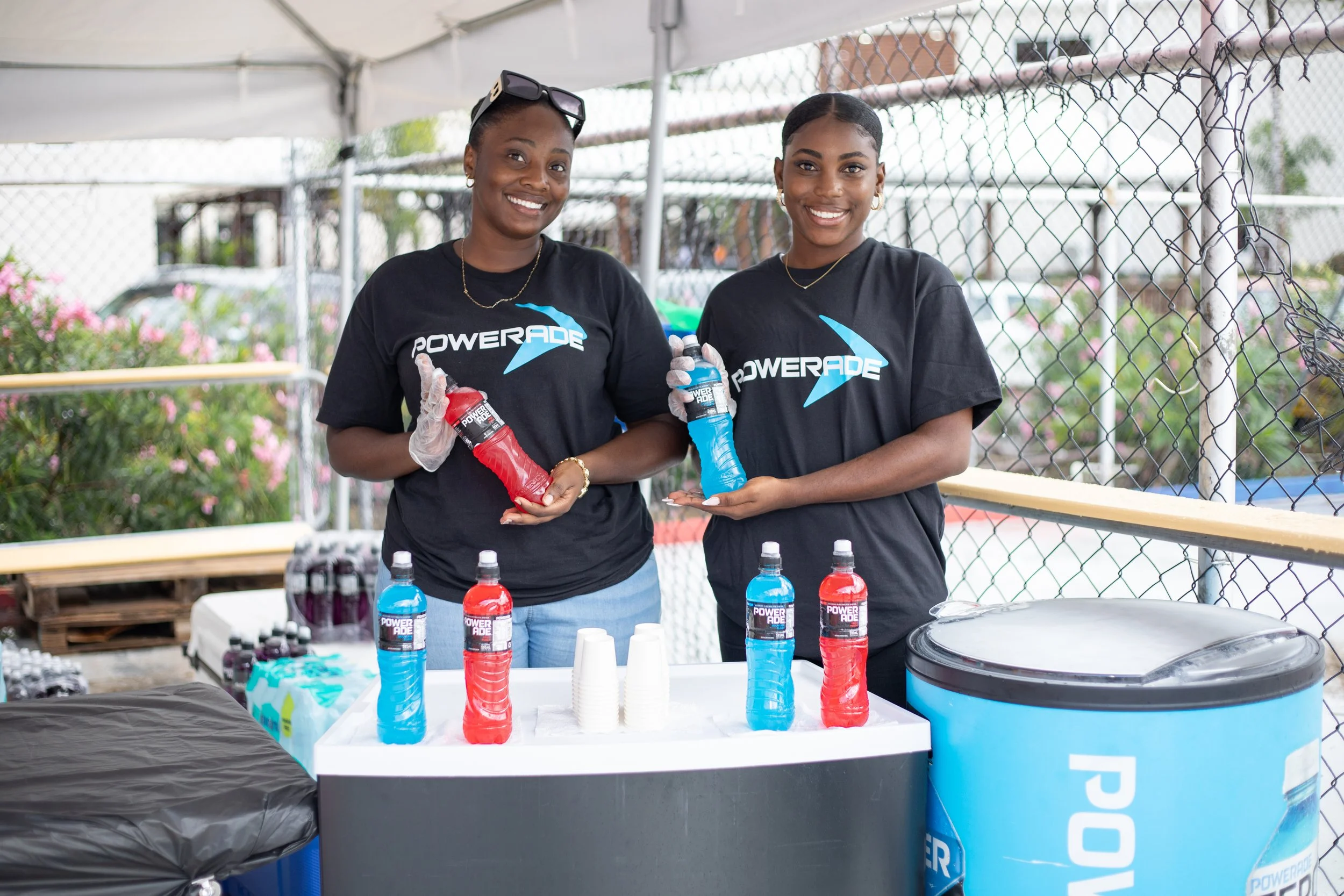 Two women wearing black Powerade t-shirts and gloves, standing behind a table at an outdoor event, holding bottles of red and blue Powerade drinks, with more bottles and cups on the table.