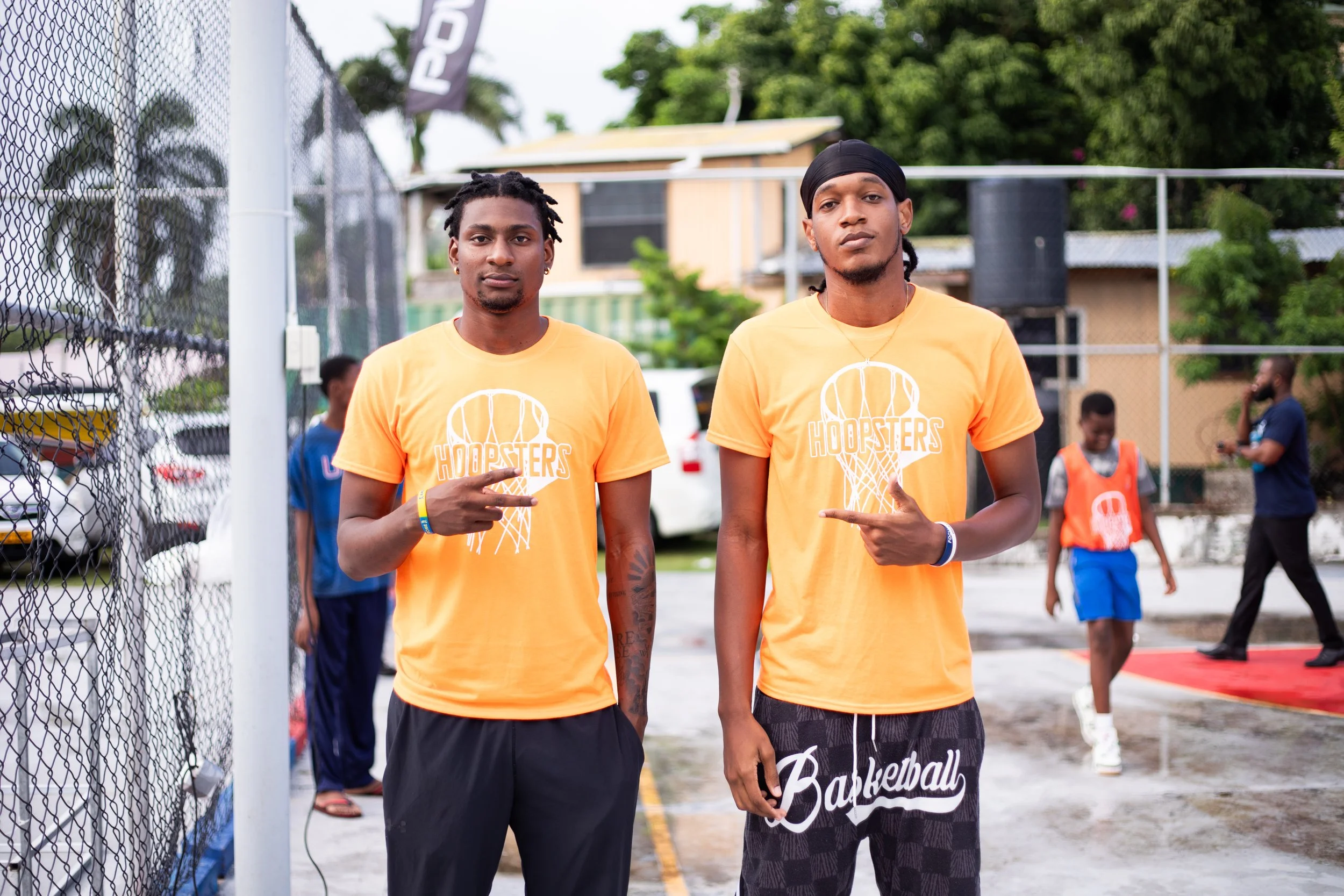 Two young men wearing orange 'Hoopsters' t-shirts posing on an outdoor basketball court, with several people in the background.