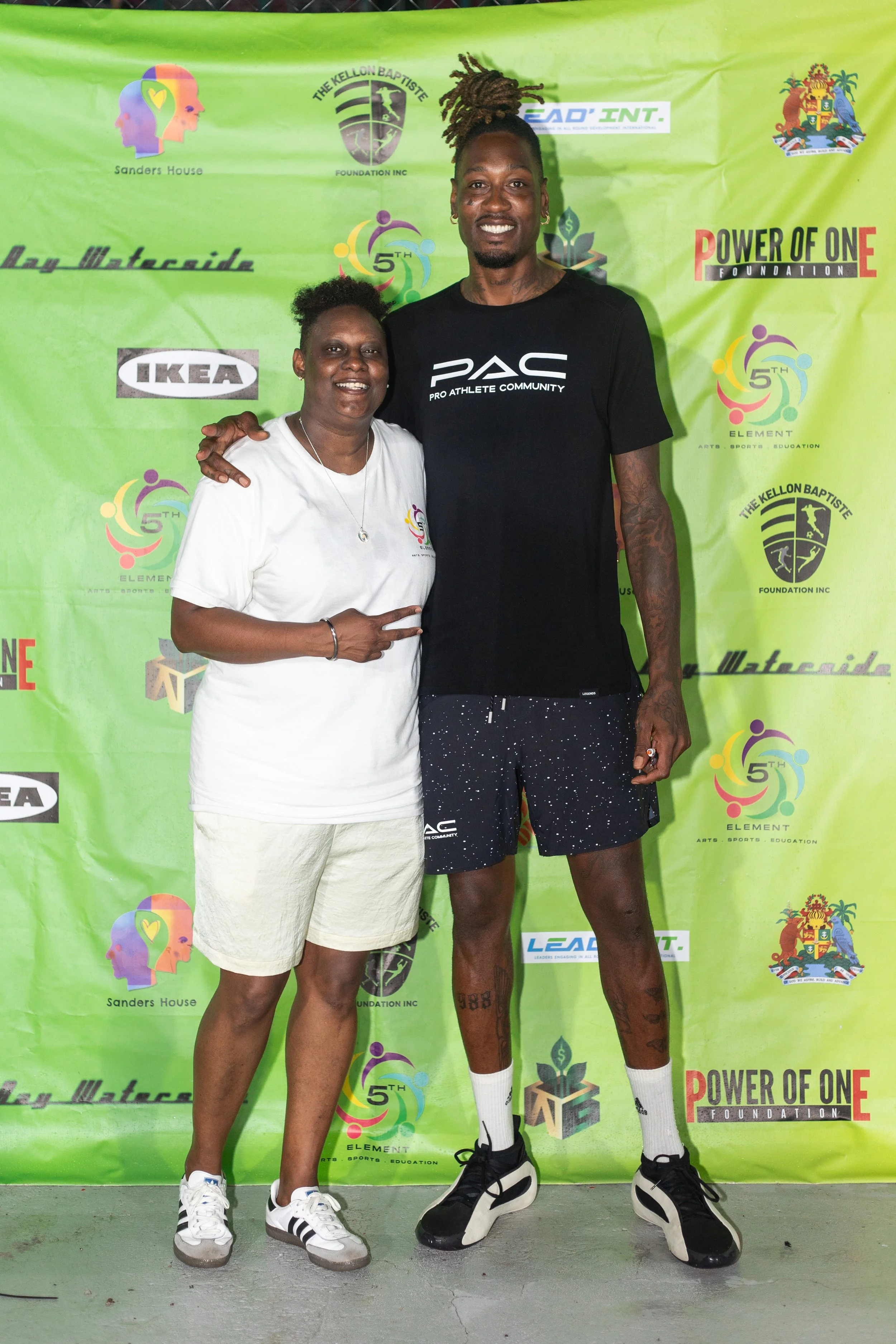 A woman and a man standing together smiling in front of a green backdrop covered with logos and text, with the woman making a peace sign and the man with his arm around her.