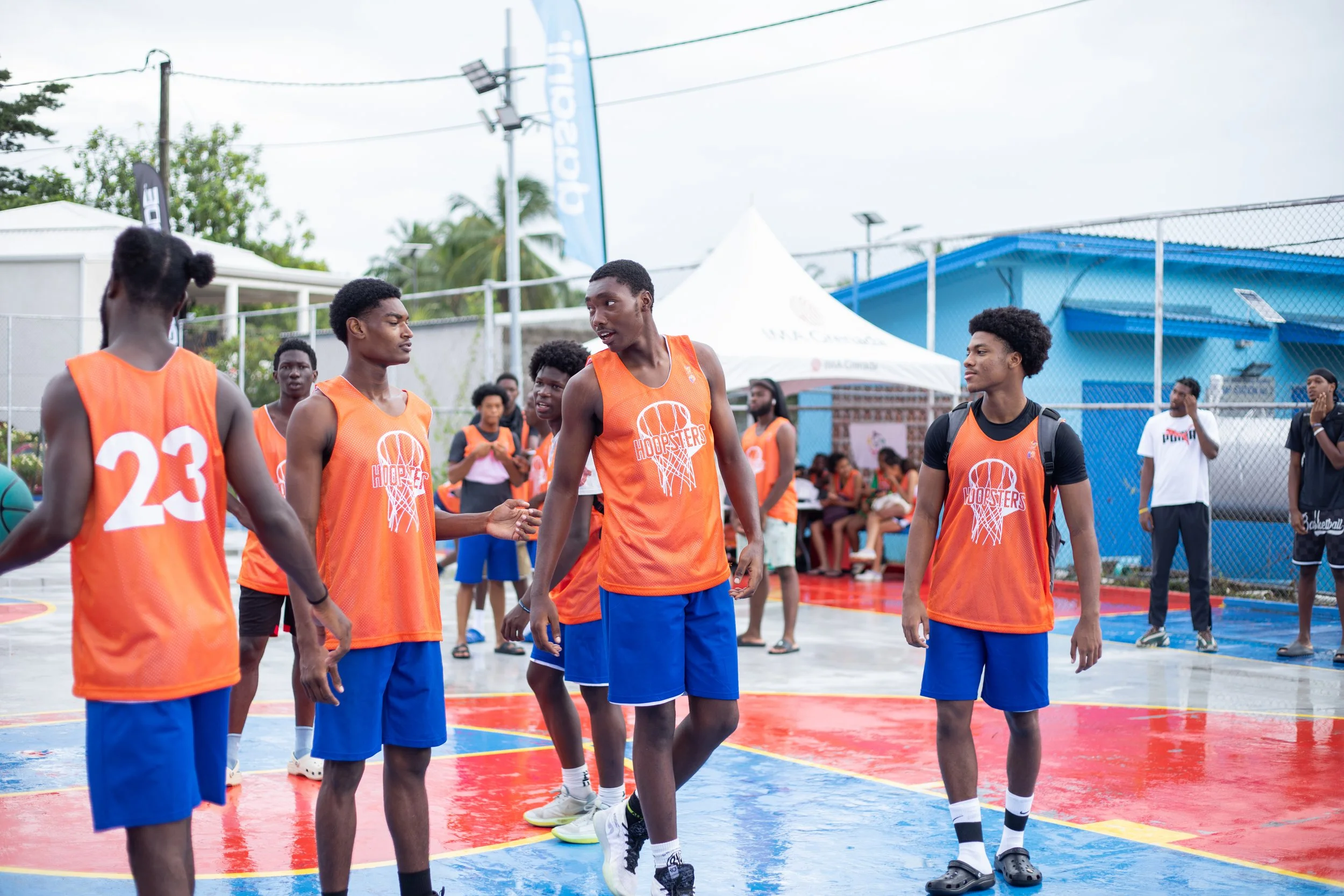 Group of young male basketball players wearing orange jerseys and blue shorts on an outdoor court, preparing for a game or practice, with some players talking and others standing around, some spectators and officials in the background.