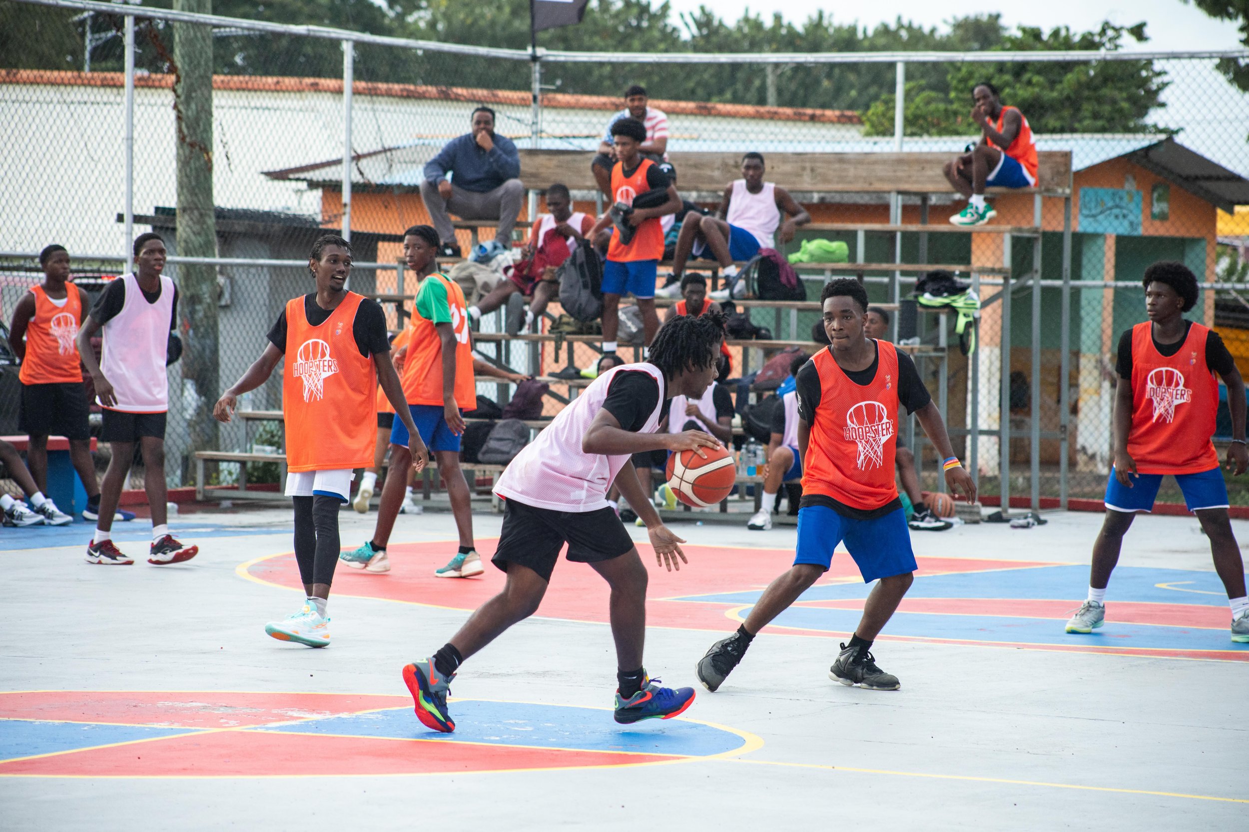 A group of young boys playing basketball on an outdoor court, wearing colorful jerseys, with some spectators sitting on bleachers in the background.