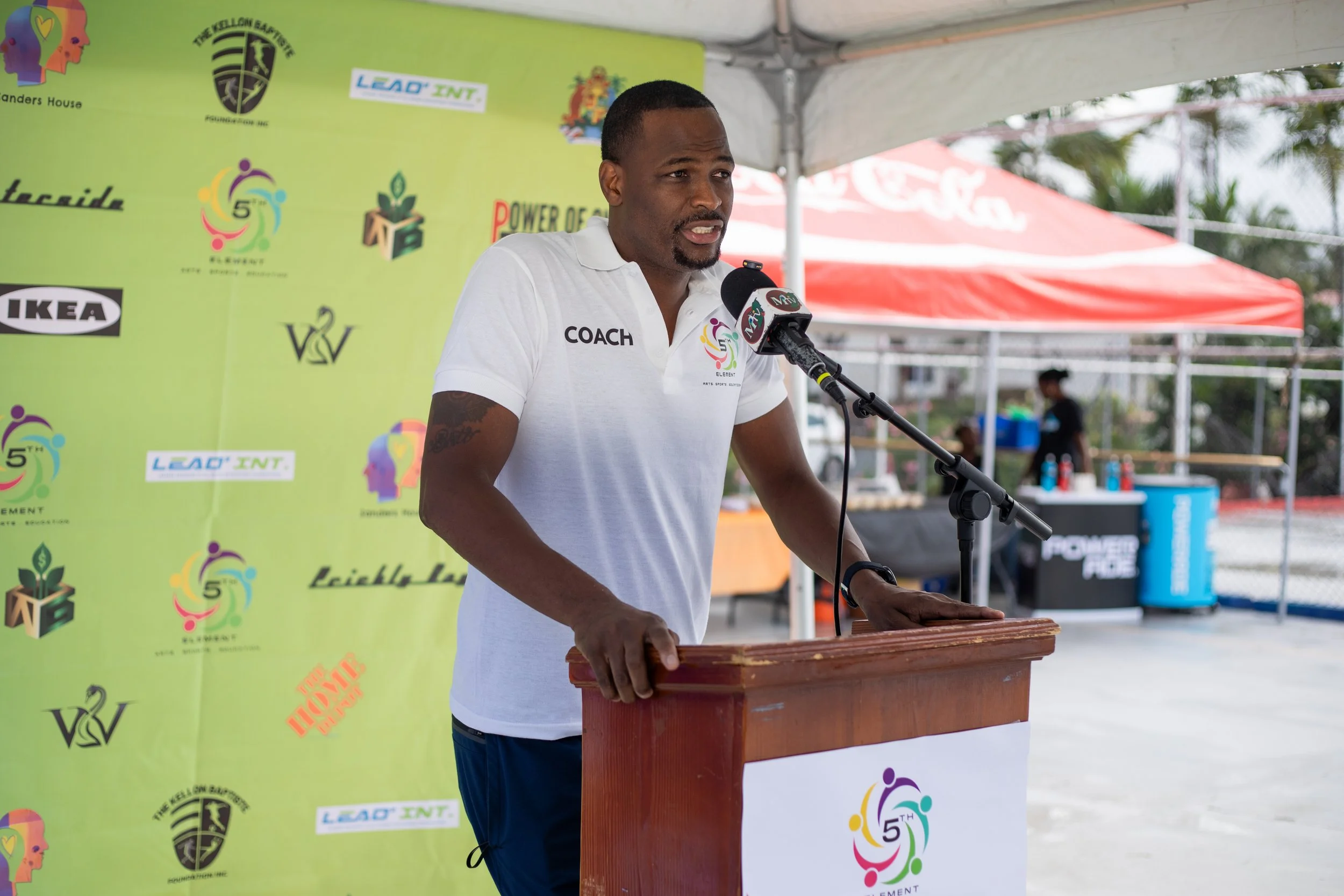 A man in a white polo shirt with 'COACH' on it speaking at a podium with a microphone, with a green backdrop and a red canopy in the background.