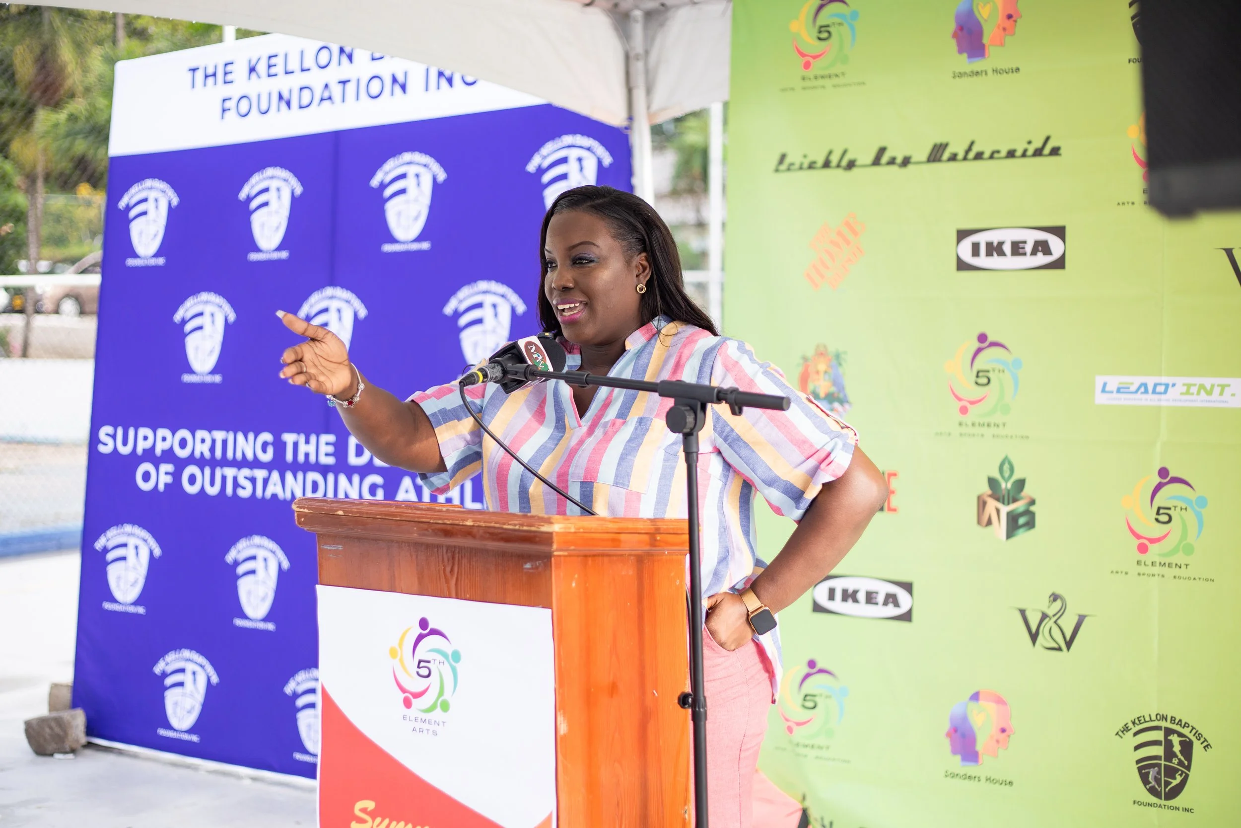 A woman in a striped shirt stands behind a wooden podium, speaking into a microphone at an outdoor event. She gestures with her left hand and has a cheerful expression. A bright blue backdrop with logos and the words supporting the development of out