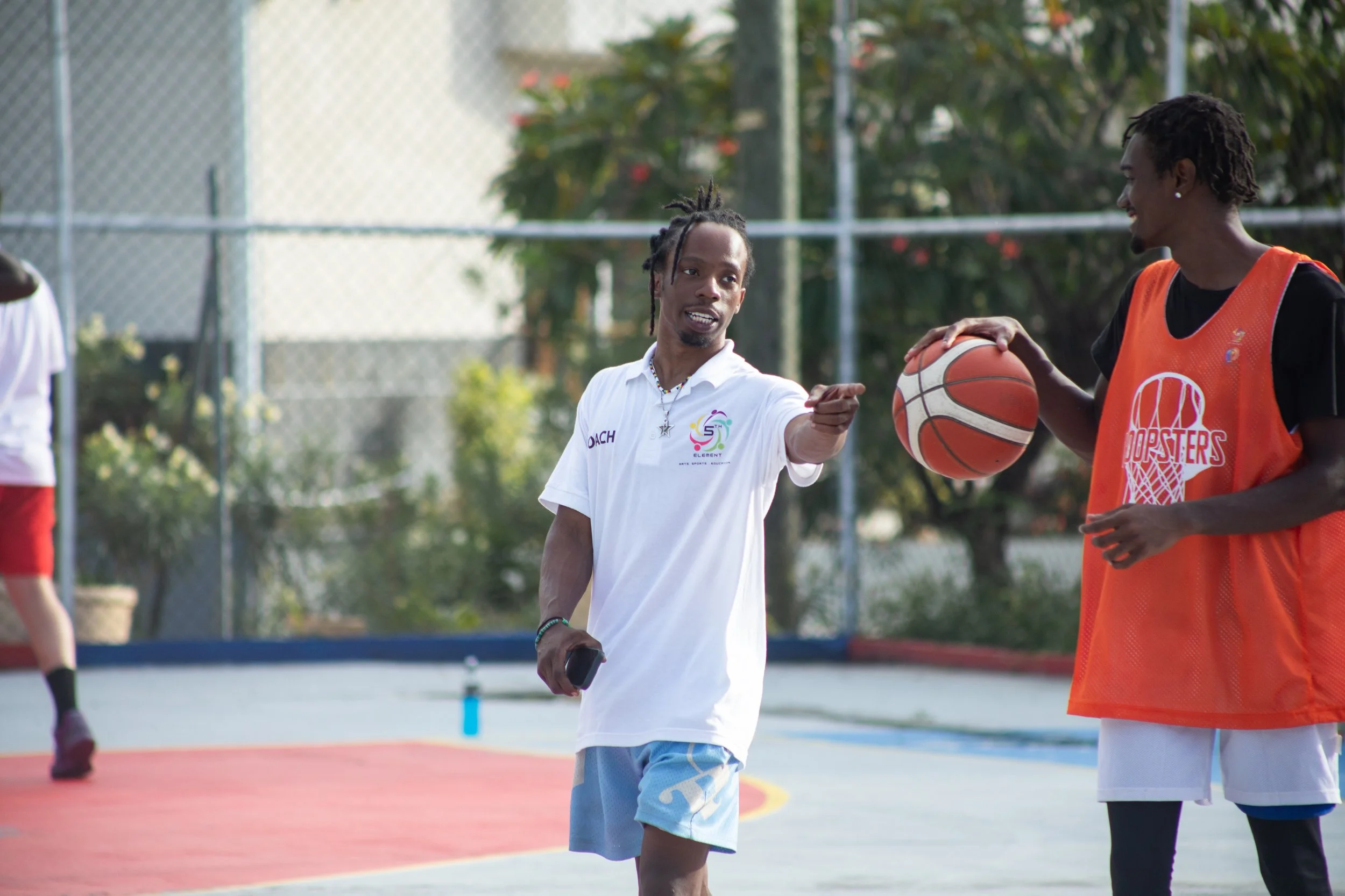 Two young men playing basketball on an outdoor court, one wearing a white shirt and the other wearing an orange jersey, with trees and a chain-link fence in the background.