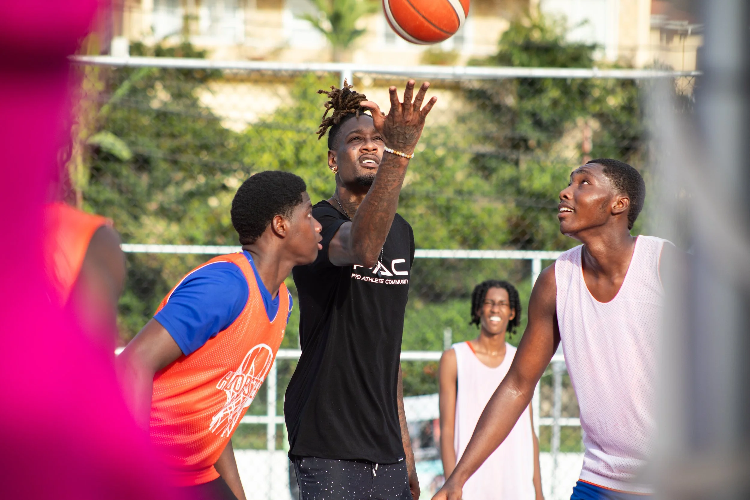 A group of young men playing basketball outdoors on a sunny day, with a player in a black shirt in the center about to shoot the ball, while two others in white and orange jerseys look on, and a woman in a pink dress in the background.