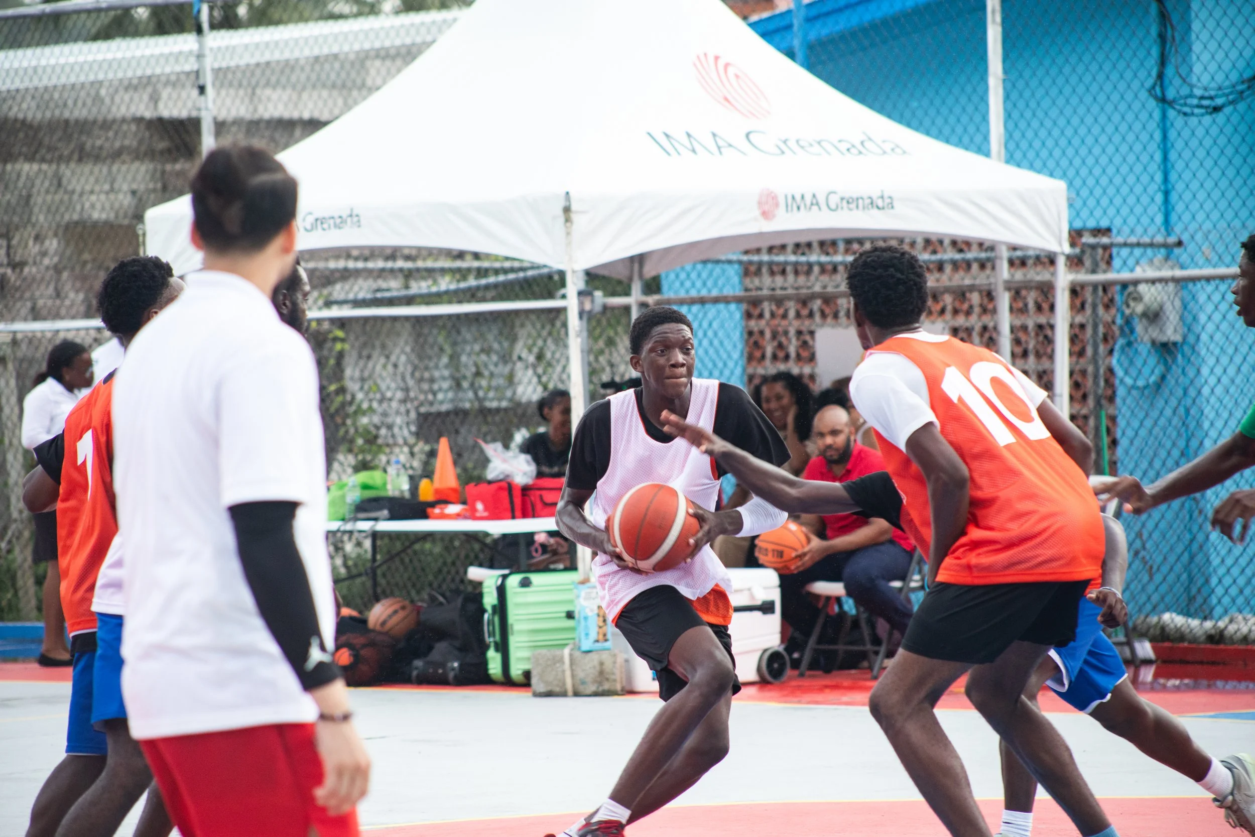 Young men playing outdoor basketball on a court, with one in a black and white jersey holding the ball, guarded by others in orange and white jerseys. A tent with 'IMA Grenada' is in the background, along with a table with equipment and spectators.