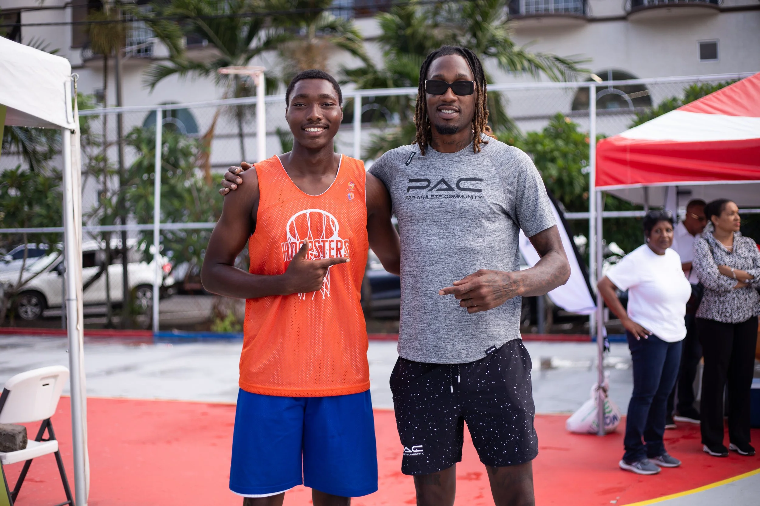 Two men posing together on an outdoor basketball court, one wearing an orange basketball jersey and the other in a gray T-shirt. The man in the jersey is smiling and pointing at himself, while the other man has his arm around him. There are people an